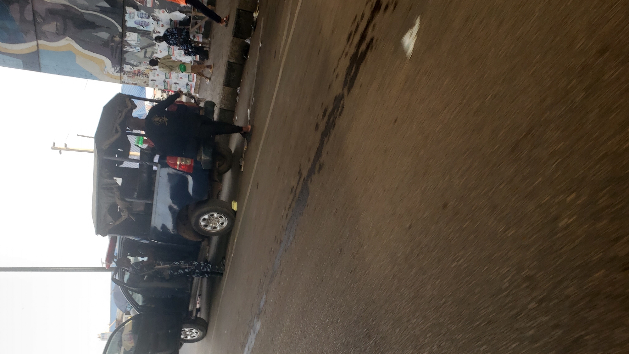The image depicts a scene in Kawo, Nigeria. A blue truck with an open back is parked on the side of the road. Several people are standing near the truck, including individuals in what appears to be police or security uniforms, one of whom is holding a rifle. The setting is an urban environment with a street running alongside a pedestrian area and a building or structure with posters/artwork. The road surface shows signs of wear and some kind of liquid staining. The photo is taken at an angle, with the horizon line tilted.