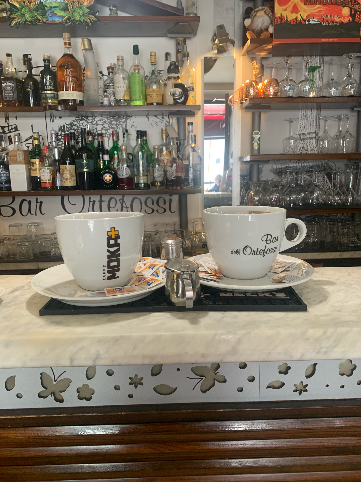The image shows a close-up view of two white coffee cups and saucers on a marble-topped bar in Abbadia San Salvatore, Italy. The cups are positioned side-by-side, with a black coaster under each. The cup on the left has the text "CAFFÈ MOKA+" vertically on its side, while the cup on the right has "Bar dell' Ortefoss" written in script. Several small packets of sugar or sweetener are scattered on the saucers. To the left of the cups, a small metal creamer or sugar dispenser is visible. Behind the cups, a shelf is lined with various bottles of liquor and other spirits, suggesting this is a bar or cafe. The background also shows shelves filled with glassware, and a decorative sign with Italian text, "Bar Ortofossi." The foreground of the image features a decorative border with cutout butterfly and floral designs. The overall environment appears to be indoors, with warm lighting.