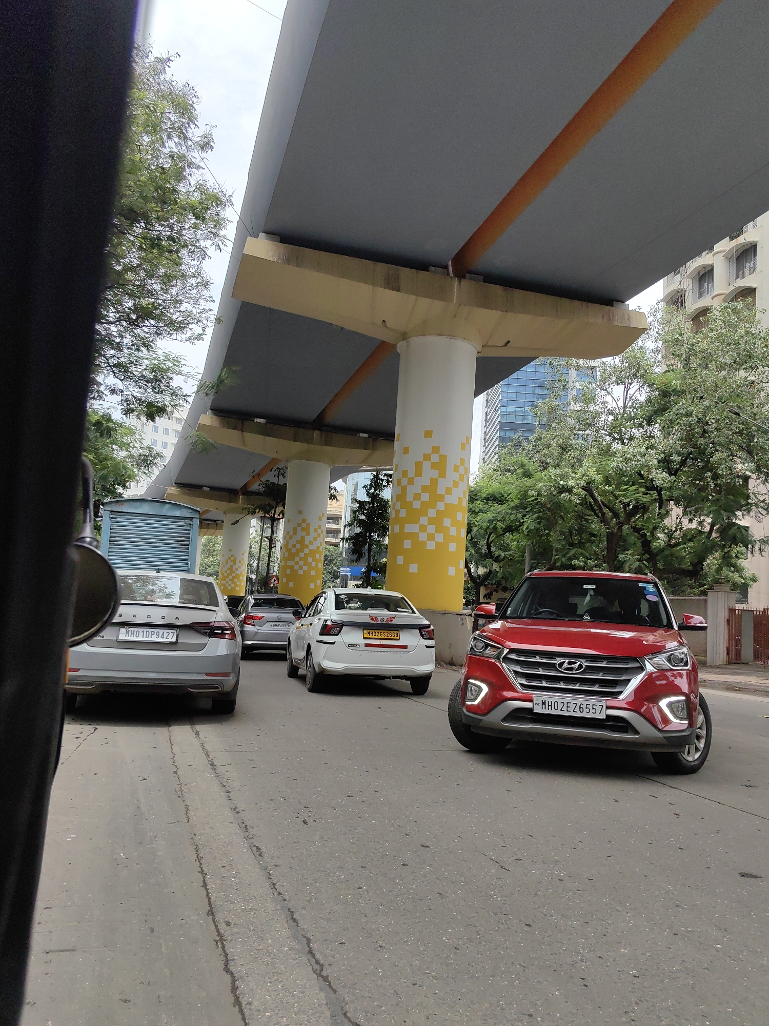 The image depicts a street scene in Mumbai, India, likely during daytime, featuring a traffic jam under a section of an elevated road or flyover. In the foreground, a red Hyundai Creta SUV is prominently positioned, its front grille and headlights clearly visible. To its left, a silver Skoda sedan with the license plate "MH01DP9427" is also stopped. Further back, a white sedan, possibly a Toyota Etios, with license plate "MH02GS2668" is visible, along with another grey sedan behind it. The elevated structure above is supported by large white pillars decorated with a yellow and white checkered pattern. Trees and urban buildings, including a high-rise structure, form the backdrop. The road surface shows tire marks and some wear. The perspective suggests the photo was taken from inside a vehicle.