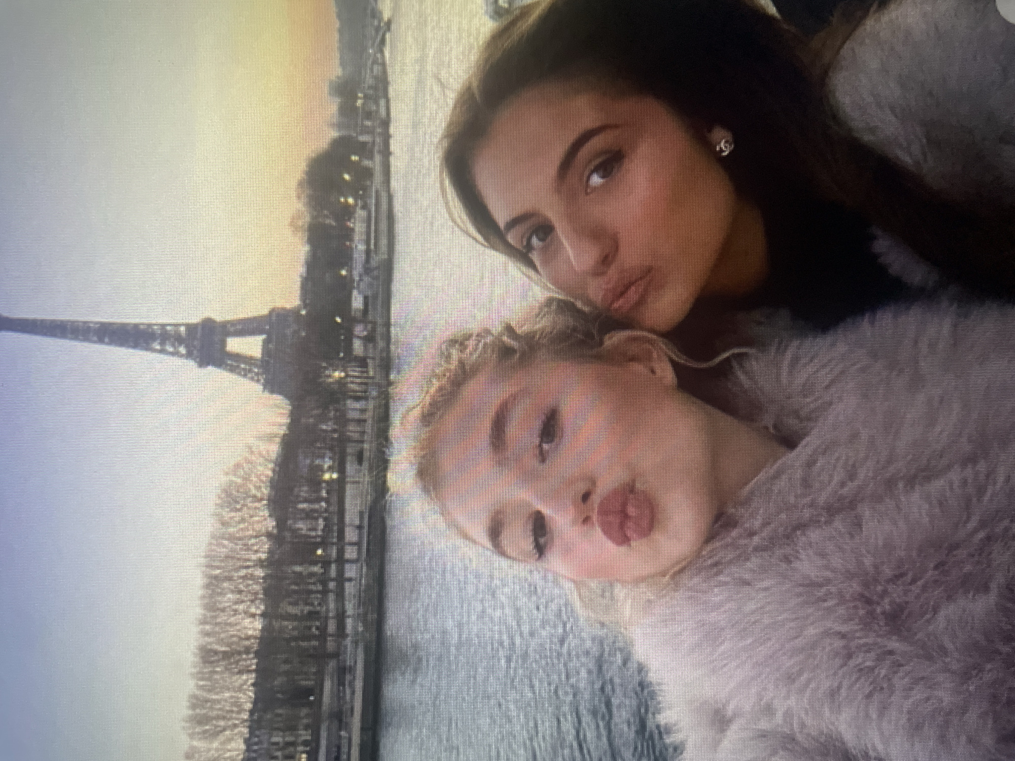 The image shows a selfie of two young women in front of the Eiffel Tower. The woman on the left has blonde hair and is wearing a fuzzy pink coat. She is puckering her lips for the camera. The woman on the right has dark hair and is wearing a black turtleneck and a gray coat. The Eiffel Tower is visible in the background, with the Seine River in the foreground. The sky is a gradient of blue and orange, indicating either sunrise or sunset. The image is of a tourist attraction in Paris, France. There is no indication of the city of Kołobrzeg, Poland.