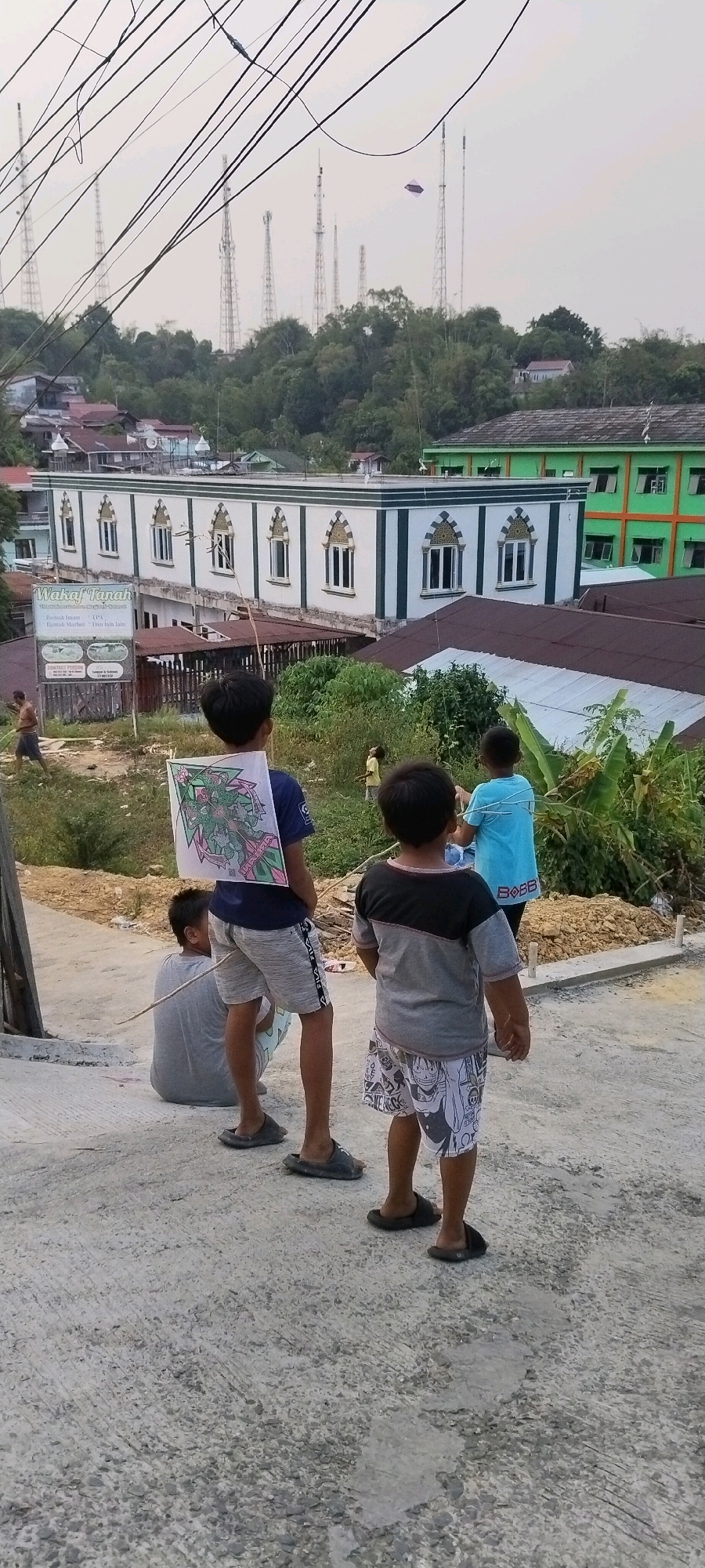 In the city of Balikpapan, Indonesia, an image captured from an elevated perspective showcases four young boys standing on a sloped, paved surface. The background reveals a densely populated area with buildings, including a mosque and a green building with orange trim, nestled amongst lush vegetation. Numerous communication towers are visible on the distant hills under a hazy sky, and a kite is flying in the upper right corner of the frame.
The boy closest to the camera, facing away, wears a grey t-shirt with a black yoke and patterned white shorts. To his left and slightly behind, another boy stands facing away, wearing a navy blue t-shirt with a large, colorful drawing of a dragon on his back and grey shorts. To the far left, a boy sits on the paved surface, facing away and wearing a grey t-shirt and shorts. A fourth boy, also facing away, is positioned to the right of the boy in the navy t-shirt, wearing a light blue t-shirt and dark shorts. The scene appears to be a residential area with signs of ongoing construction or development, suggested by piles of dirt and materials in the mid-ground.