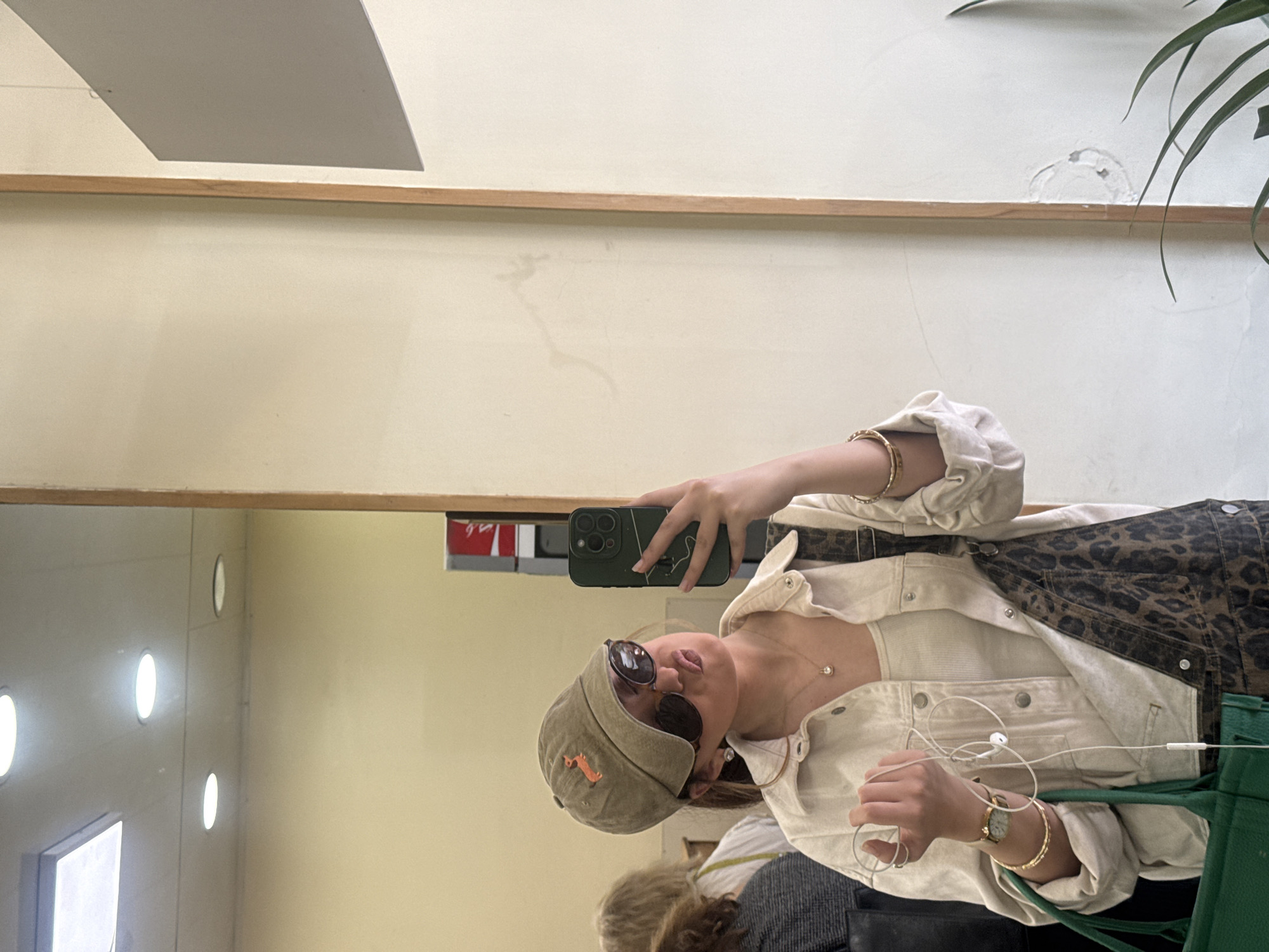 A woman is taking a selfie in a mirror. She is wearing a beige baseball cap with a dog emblem, sunglasses, a beige jacket, and jewelry. She is holding a dark green cell phone in her right hand and tangled white wired headphones in her left. In the reflection, there are other people and objects, including a green tote bag, and the interior of a brightly lit room with round lights. The top of the mirror shows part of a white wall with a wooden trim. The location is possibly in Yerevan, Armenia.