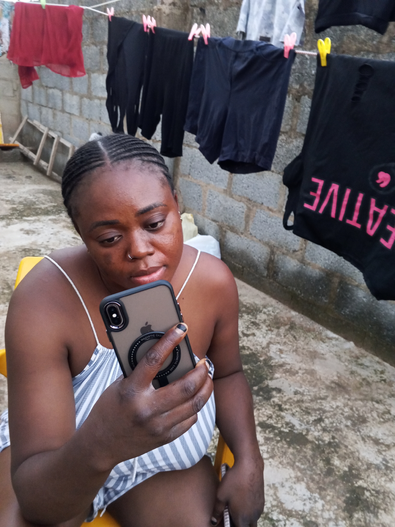 An outdoor, eye-level shot captures a woman sitting on a yellow chair, engrossed in her smartphone. She has braided hair, wears a nose ring, and is dressed in a white and grey/blue striped spaghetti-strap garment. Her fingers, adorned with dark nail polish, hold a brown smartphone with a clear case that reads "MAGNETIC CASE" and features a circular magnetic ring on its back. Her expression is focused as she looks down at the screen.
In the background, clothes are drying on a line against a rough concrete block wall. From left to right on the clothesline, a red sheer garment hangs, followed by several black garments resembling pants or leggings. A light grey top and black shorts are also visible. On the far right, a black top or t-shirt with pink lettering that partially reads "EATIVE" and a pink speech bubble icon is hanging. Clothespins in red, pink, and yellow secure the laundry. Below the clothesline to the left, a wooden ladder or frame leans against the wall. The ground beneath is a textured concrete or paved surface. The setting appears to be a typical outdoor residential area in Abuja, Nigeria, during the daytime with diffused light.