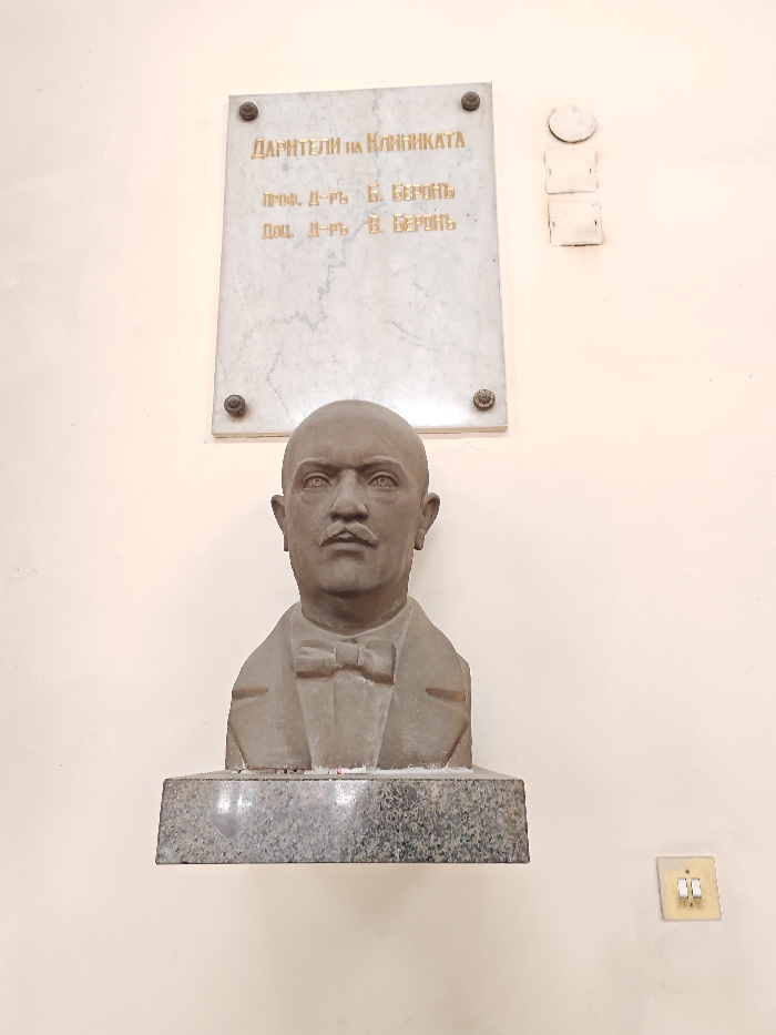 An indoor, eye-level photograph captures a commemorative bust and an informational plaque mounted on a plain, light beige wall. The scene is located within a medical facility, specifically a clinic or hospital, in Sofia, Bulgaria. Centrally positioned, a grey, patinated bust depicts a bald, mustachioed male figure with a resolute expression, wearing a suit jacket and a bow tie. The bust rests on a dark, speckled granite base. Directly above the bust, a rectangular white marble plaque, secured by four bronze-colored screws, displays gilded Cyrillic text. The inscription reads: "ДАРИТЕЛИ НА КЛИНИКАТА" (DONORS OF THE CLINIC), followed by "ПРОФ. Д-РЪ Б. ВЕРОНЪ" (PROF. DR. B. VEROV) and "ДОЦ. Д-РЪ В. ВЕРОНЪ" (ASSOC. PROF. DR. V. VEROV). To the upper right of the plaque, three small, off-white, rectangular electrical covers are stacked vertically. In the lower right corner of the image, a cream-colored double-toggle light switch is affixed to the wall.