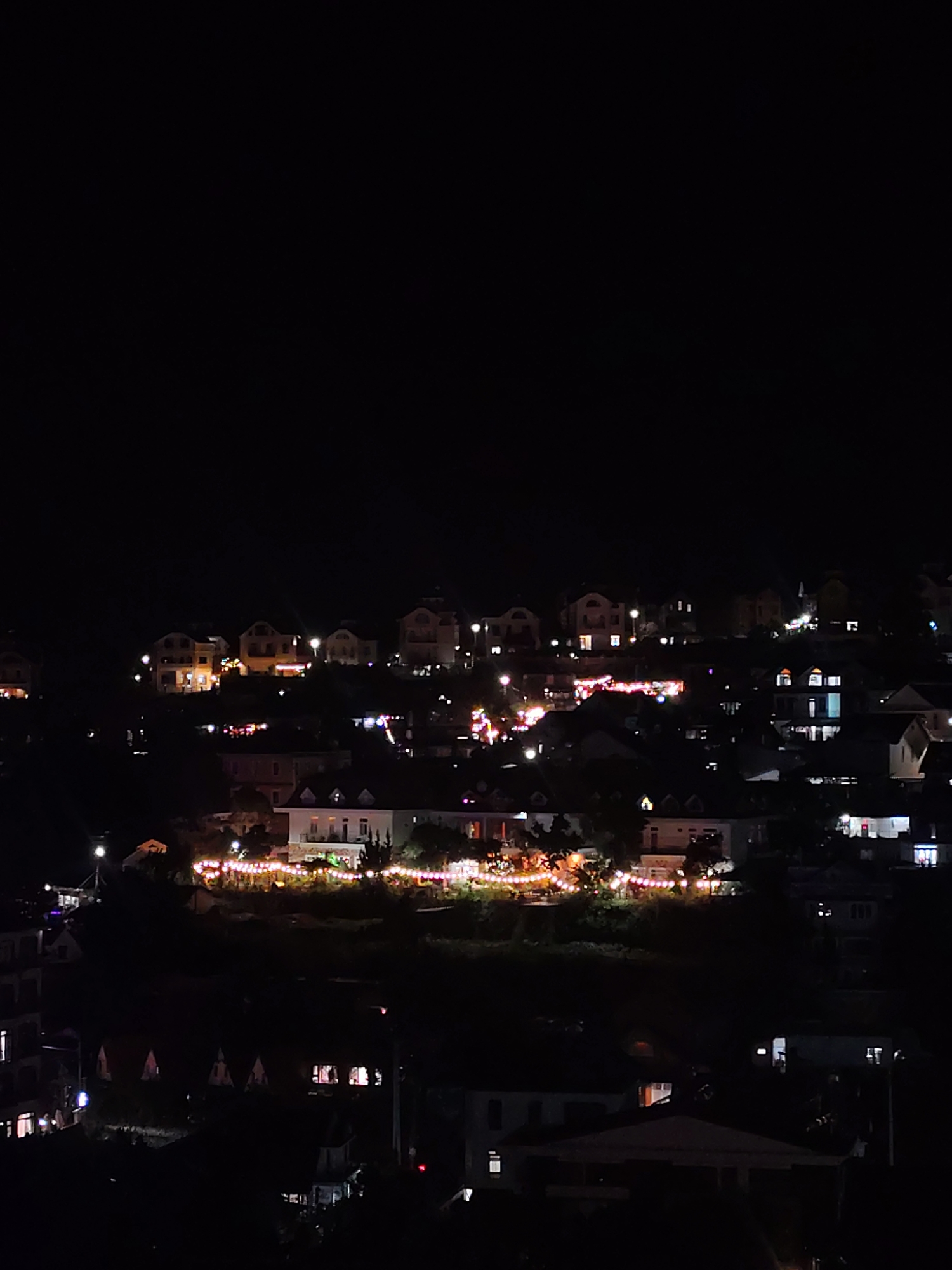 The image captures a night scene of a hillside residential area in Da Lat, Vietnam, characterized by a dark sky and the illumination of numerous houses and buildings. The structures are nestled on varying elevations, creating a tiered effect across the landscape. Many of the houses are lit from within, casting warm yellow and orange glows through their windows. Additionally, decorative string lights are visible in several areas, particularly along the edges of some properties and what appear to be outdoor seating or dining areas. These lights add a festive and ambient quality to the scene. The overall impression is of a peaceful, residential neighborhood at night, with the lights creating a sense of warmth and activity against the dark backdrop of the surrounding environment and the expansive night sky.