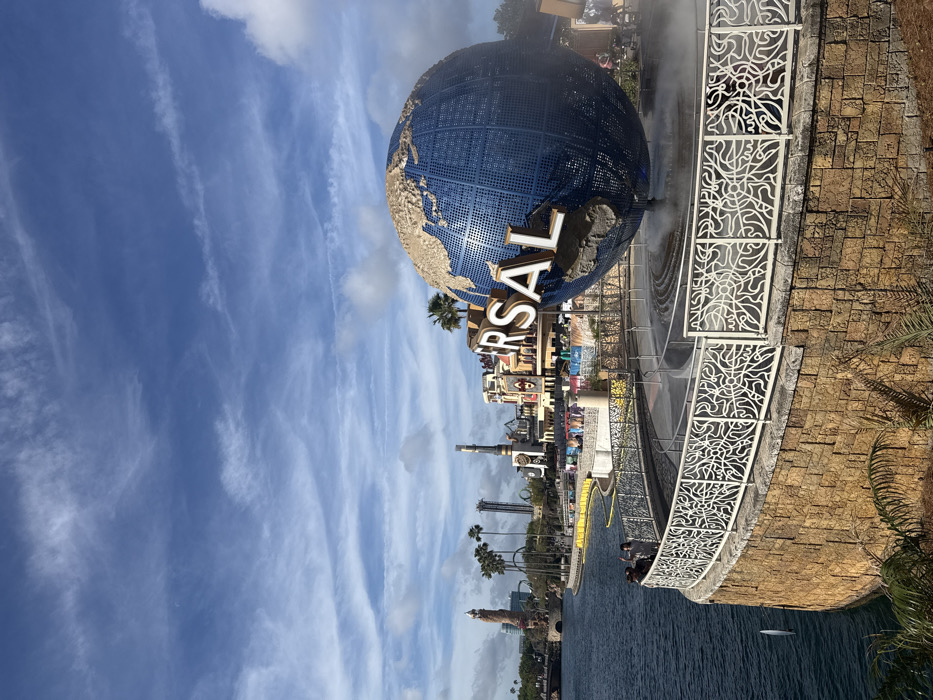This image depicts the iconic Universal Studios Globe, a large, rotating spherical sculpture of Earth, located at Universal Orlando Resort in Orlando, Florida, United States of America. The blue globe features gold-colored landmasses and is partially obscured by the white, three-dimensional letters spelling "UNIVERSAL". It is positioned within a circular water feature from which mist is actively emanating.
In the foreground, a curved retaining wall constructed of irregular tan and brown stone blocks supports a white metal railing. This railing exhibits an intricate, abstract, and organic pattern. To the left of the railing, a placid body of water extends into the background. Two individuals are visible on the walkway adjacent to the railing, facing the water; one individual is partially obscured and appears to be gesturing.
The background, across the body of water, reveals a segment of a theme park. Notable structures include a tall, light brown cylindrical tower resembling a lighthouse, multiple green palm trees, and various multi-story buildings and thrill ride structures with complex architectural details. The sky overhead is clear blue with scattered white, wispy clouds and several visible contrails. The scene is uniformly illuminated by natural daylight.