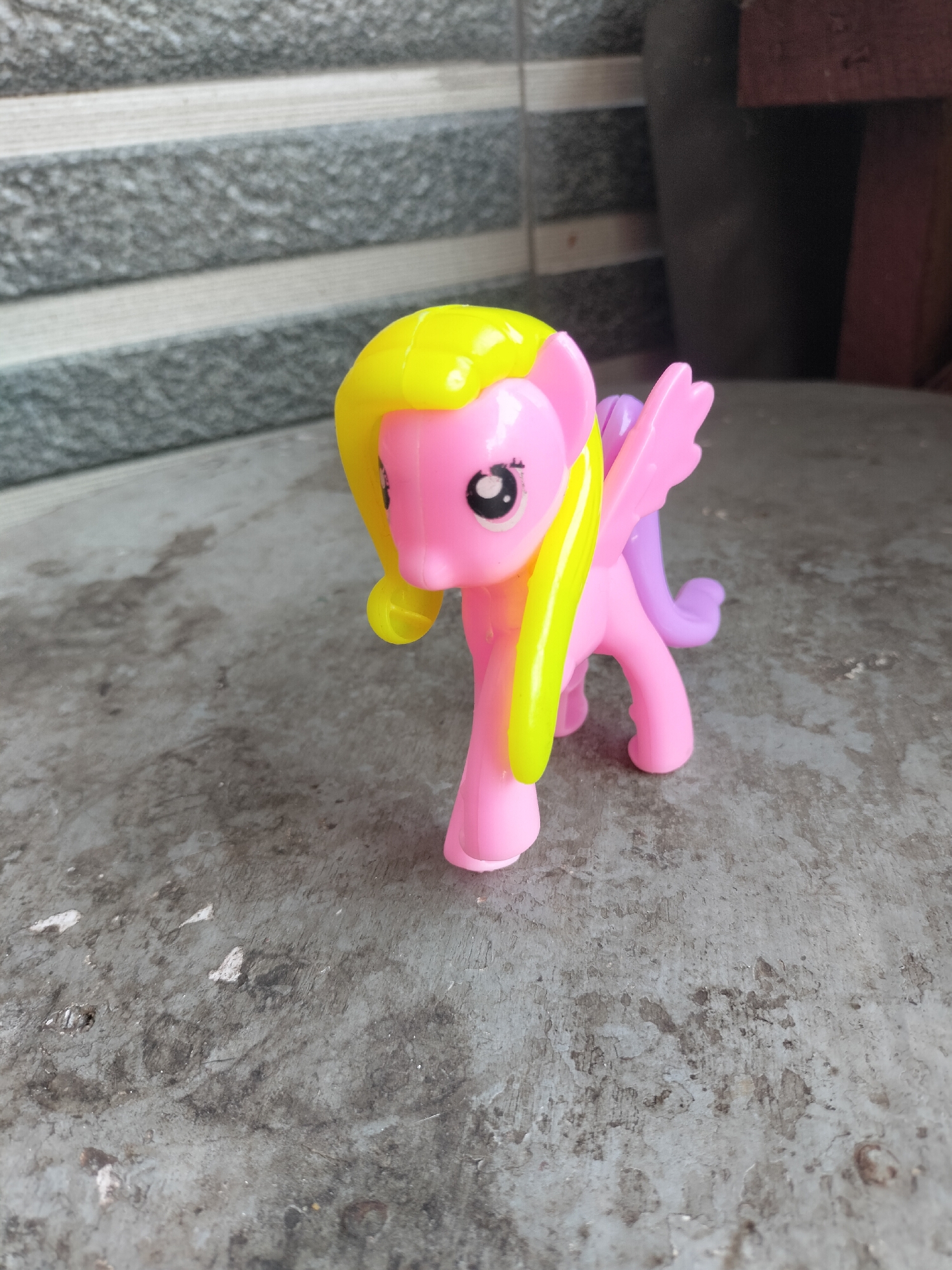 A pink toy pony with yellow hair and purple wings is standing on a concrete surface. The background consists of a textured grey wall. The scene appears to be outdoors, possibly in a courtyard or on a patio. The lighting suggests it is daytime. The image is taken from a slightly elevated angle, looking down at the toy.