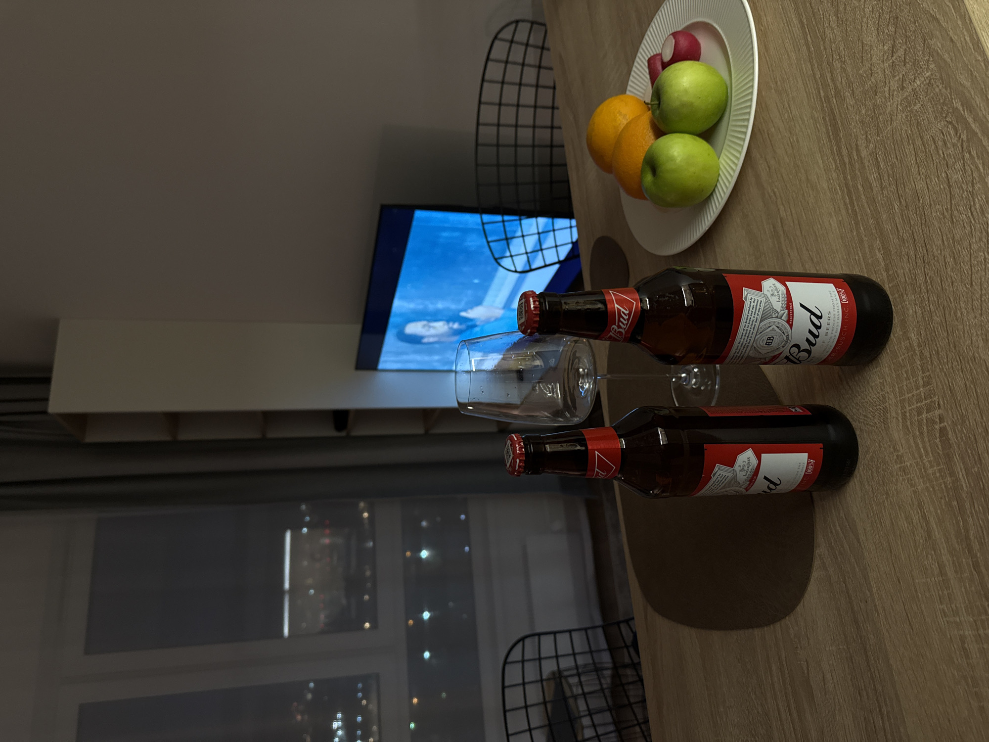 The image shows a scene indoors, likely in an apartment. On a wooden table, there is a plate of fruit including oranges, green apples, and radishes. Next to the fruit are two bottles of Budweiser beer and a wine glass. A television in the background displays a scene with blue sky and clouds. Through a window, a cityscape can be seen, suggesting an urban environment. The lack of specific identifiers makes it impossible to determine the exact location in Moscow, Russia, without additional information. There are no people in the scene.