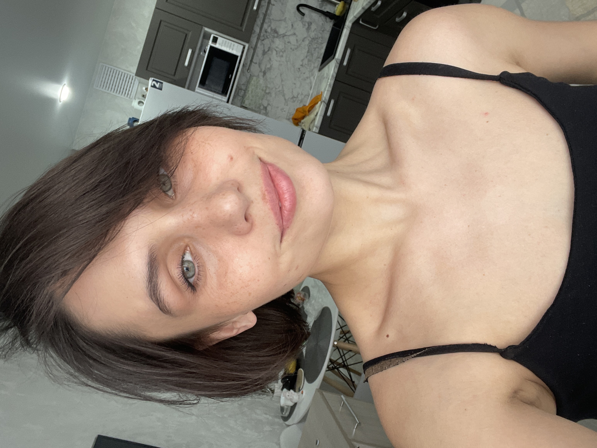 This close-up selfie, captured in Izhevsk, Russia, features a young adult female with short dark brown hair and light-colored eyes, possibly green or grey-green. She has visible freckles on her nose and cheeks and is smiling softly with her mouth closed. She wears a black tank top or camisole with thin straps; one strap near her collarbone appears to have a lace-like texture. A small reddish blemish is visible on her right shoulder (viewer's left), and a darker mole on her neck/shoulder area (viewer's right).
The woman is positioned at an angle, as the image is rotated counter-clockwise. The background reveals an indoor home setting, primarily a modern kitchen with dark grey cabinets, both upper and lower, and a light-colored countertop, possibly marble. A stainless steel microwave oven is integrated into the cabinetry, and a dark kitchen faucet is visible. Overhead, a ceiling light illuminates the scene.
Further in the background, a white refrigerator with a sticker displaying the number "2" is partially visible, along with a roll of paper towels. Near the floor, a white and black robot vacuum cleaner is seen, as well as part of a modern chair. The overall lighting suggests an indoor setting, possibly with artificial light sources. The woman's expression conveys a relaxed and pleasant mood.