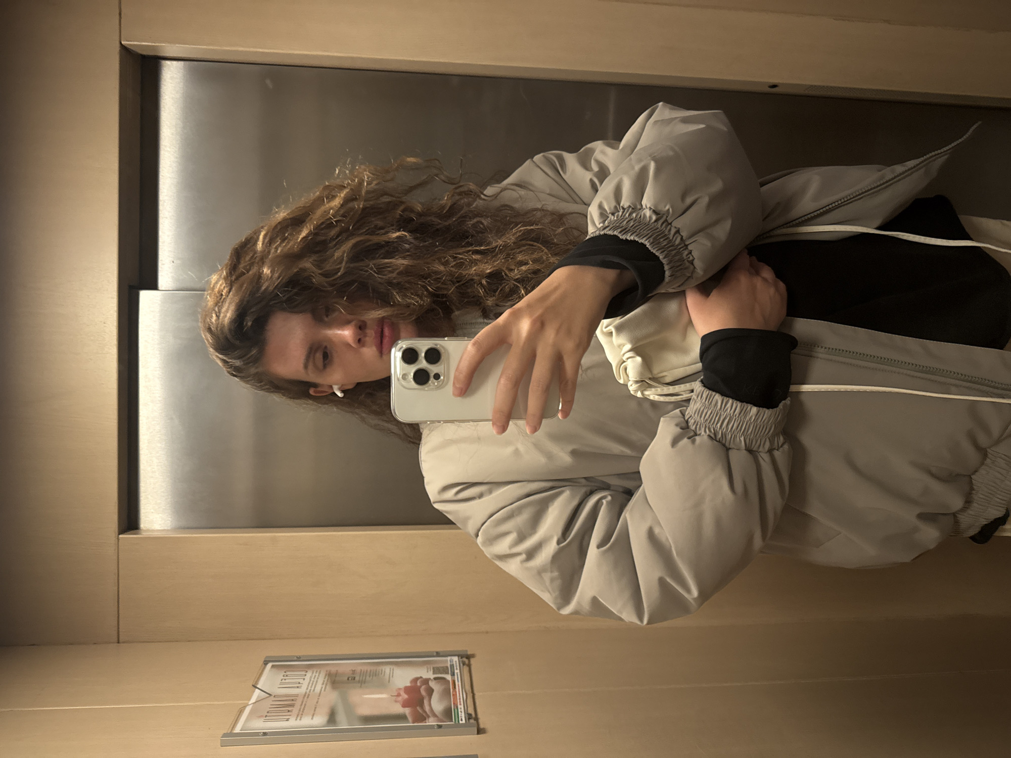 A woman with curly brown hair is taking a selfie in an elevator. She is wearing a light grey puffer jacket and a black shirt. She is holding a white iPhone in her right hand. The elevator has a stainless steel back wall and light brown wood paneling on the sides. There is a framed advertisement on the wall of the elevator, near the bottom of the shot. The text on the ad is Cyrillic. The woman's face is partially obscured by the phone. Based on the Cyrillic text on the advertisement and the architectural style of the elevator interior, the image was likely taken in Russia. Without additional information, it is difficult to confirm that the location is Krasnoyarsk, Russia.