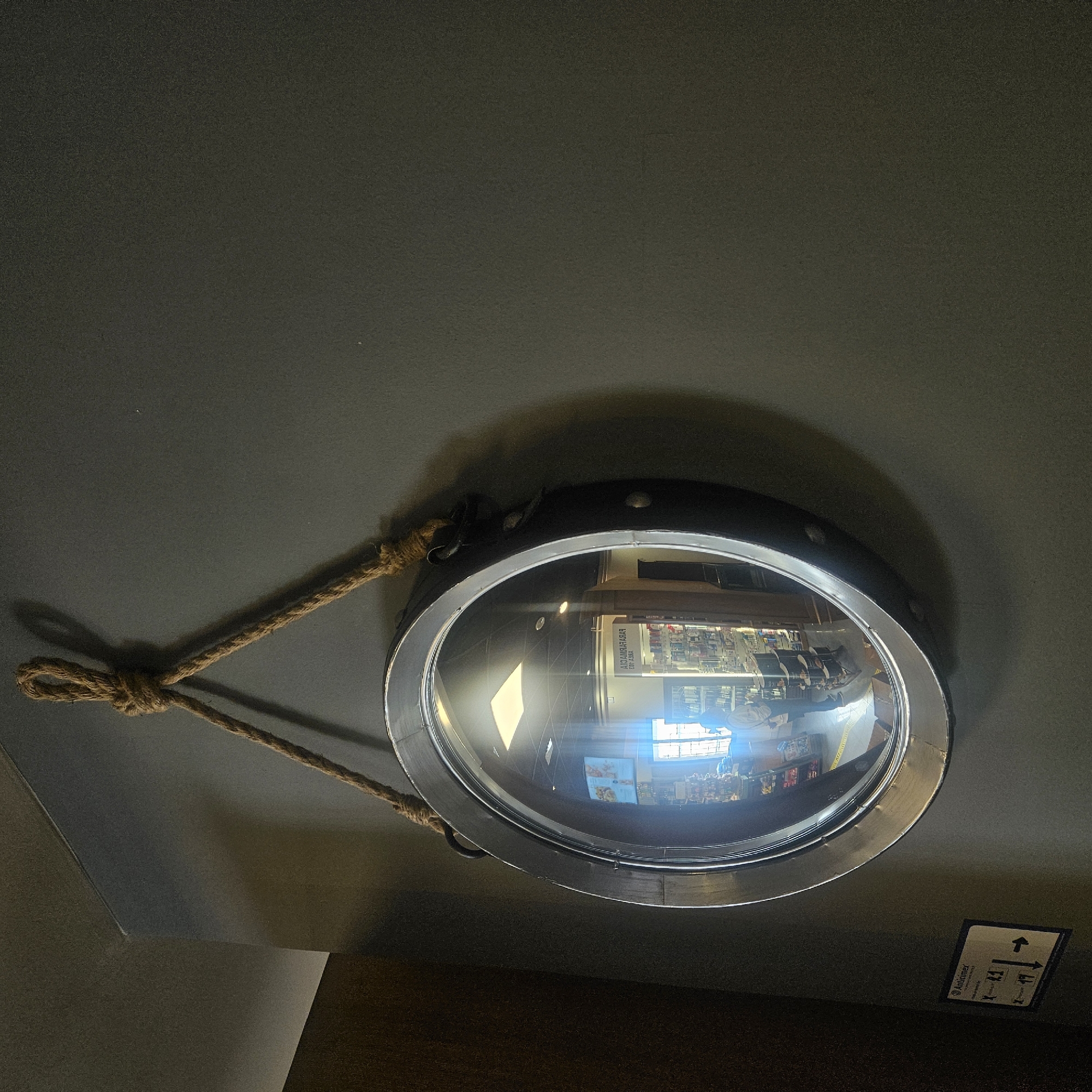 A round, nautical-style mirror with a rope hanger is affixed to a dark gray wall. The mirror's frame is metallic, with visible rivets. The reflection in the mirror shows the interior of a shop, possibly a pharmacy, with shelves stocked with products, fluorescent lighting, and a person standing and facing away from the camera. A sign with the text "Anticimex" and directional arrows is also visible on the wall to the left of the mirror. The overall scene suggests an indoor environment. This scene is not located in Almadrones, Spain.