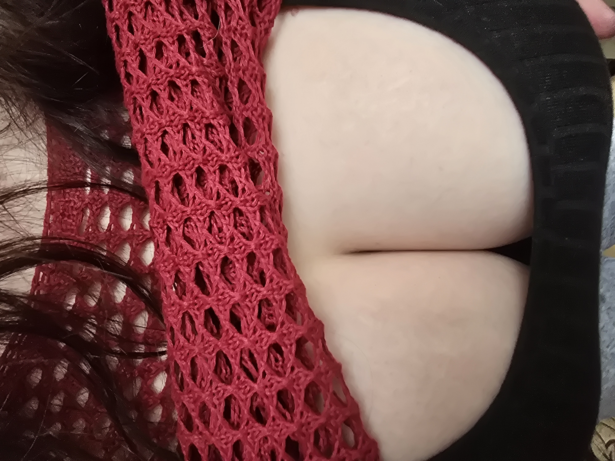The image shows a close-up, overhead view of a person's chest. The person has light-colored skin and dark hair. They are wearing a red, crocheted, open-weave top, and a black garment, possibly a sweater or dress. The red top is draped over the chest, revealing a cleavage area. The lighting is bright and appears to be coming from above and slightly to the left. The background is out of focus but appears to be a neutral, indoor setting. No specific time of day, weather, or location cues are discernible. There is no visible text in the image. The composition focuses on the texture of the clothing and the skin, with a shallow depth of field.