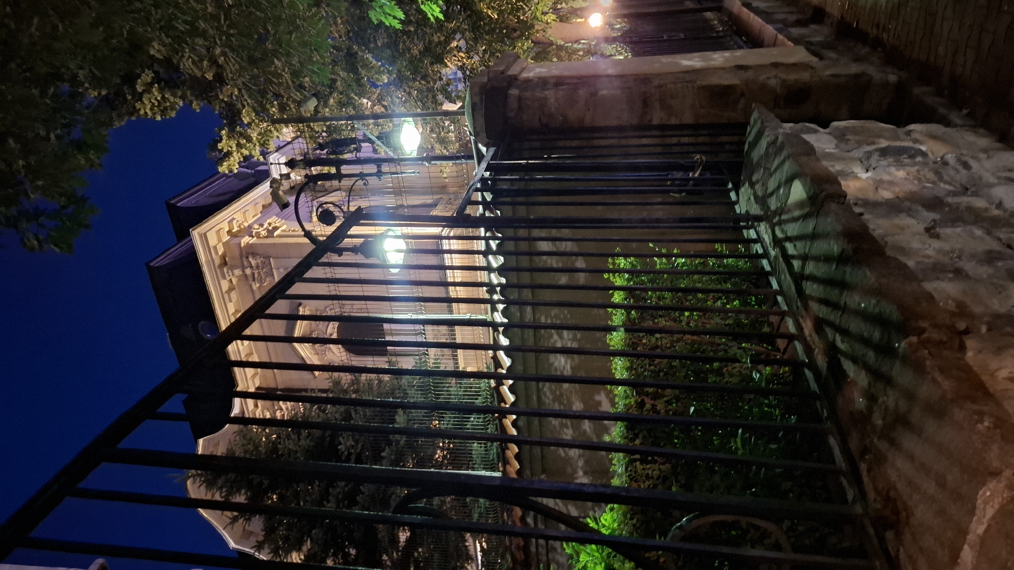 The image captures a dimly lit, imposing stone wall and a black metal fence in the foreground, with a grand building visible behind it at night. The building appears to be a multi-story structure with ornate architectural details, illuminated by warm lights, casting long shadows from the fence onto the stone wall and ground. A lush green hedge runs along the base of the fence. Two vintage-style lamps are mounted on the fence posts, emitting a soft green glow. Trees with foliage are visible to the right of the building, with another light source visible further down the street. The sky is a deep, dark blue, indicating nighttime. The overall scene suggests a property of historical or significant architectural value in a European city. This image was taken in Sofia, Bulgaria, specifically in front of the "House with the Lime Tree" (Къщата с липите), located at 17 Tsar Ivan Shishman Street.