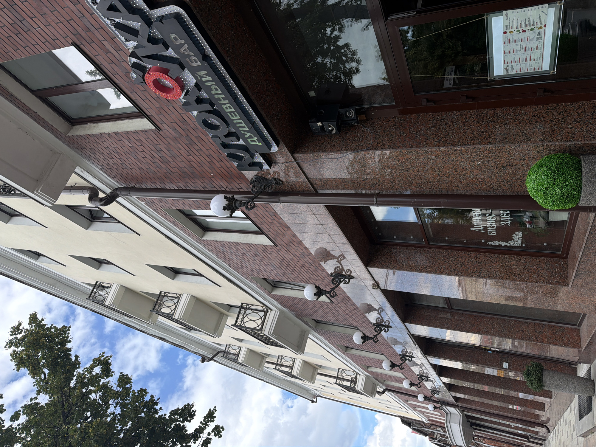 The image shows the exterior of a building with a brick facade and several windows. A sign with dark, three-dimensional lettering and a red circular element is mounted on the brick wall. Below the sign, a series of ornate streetlights with white globes are attached to the building. The building's facade transitions to polished granite in some sections. A large potted plant with green foliage stands near the entrance. Through a glass window, a menu is visible, displaying text in a foreign language. The sky above is partly cloudy, suggesting daytime. The image is oriented vertically, with the bottom of the building appearing at the top of the frame. The text visible on the sign appears to be in Cyrillic script. The overall scene suggests a commercial establishment in an urban setting.