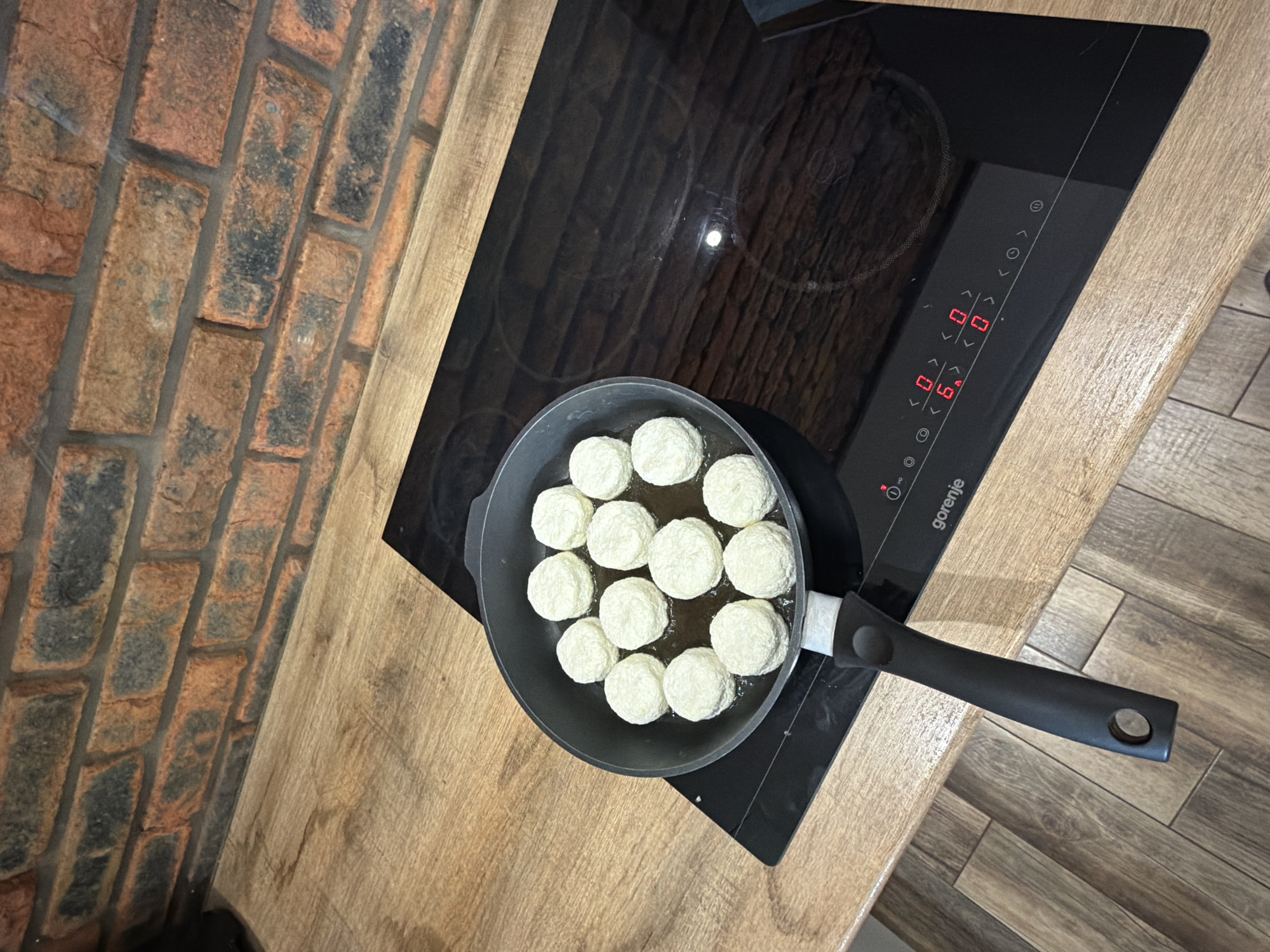 This is an eye-level shot of a kitchen scene, with the camera angled slightly downwards. A black frying pan filled with several round, white objects, possibly cottage cheese pancakes or small dumplings, rests on a black electric stove top. The stove's control panel is visible, displaying red digital numbers. To the left of the stovetop, a brick wall is visible, next to a wooden countertop. To the right, a different wooden surface is visible. The kitchen appears to be located in the city of Chaikovsky, Russia.