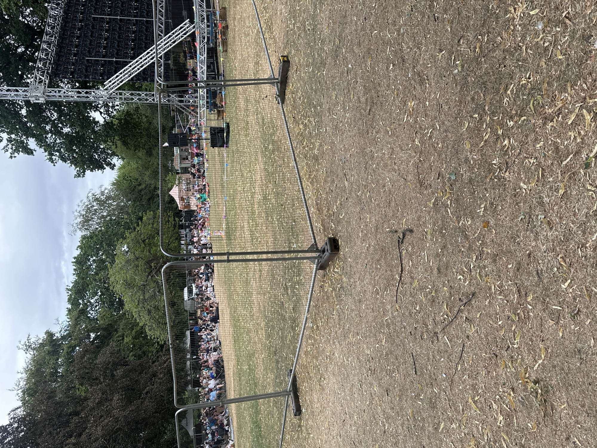 The image shows an outdoor event with a crowd gathered on a grassy field in the city of Rugby, United Kingdom. Temporary fencing separates the foreground from the audience area, where people are seated on blankets and chairs. Behind the crowd, a large stage structure with a screen is visible, along with tents and trees. The ground in the foreground is dry and covered with fallen leaves. The sky is overcast.