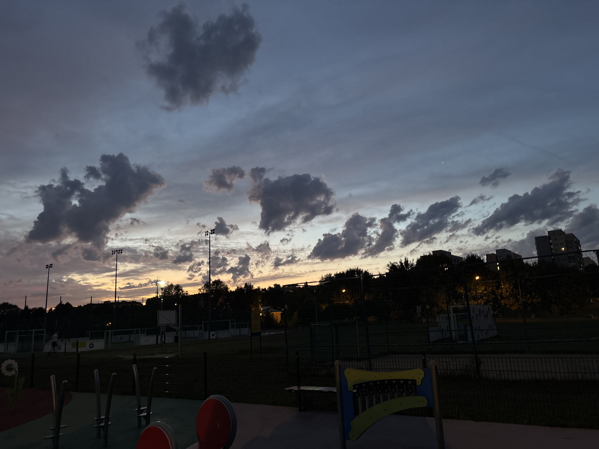 The image depicts a sports field at dusk, likely in a residential area of Lublin, Poland. The sky is a gradient of dark blue and grey, with scattered clouds, and a hint of orange near the horizon from the setting sun. Tall light poles with multiple lights are visible on the field, some of which are illuminated, casting a yellowish glow. The field itself appears to be a multi-purpose sports area, with fencing and markings visible. In the foreground, a colorful playground structure with metal bars and red circular elements is partially visible. Behind the field, trees and buildings, including multi-story apartment blocks, can be seen silhouetted against the twilight sky. The scene is calm and quiet, suggesting it is either early evening or late evening.