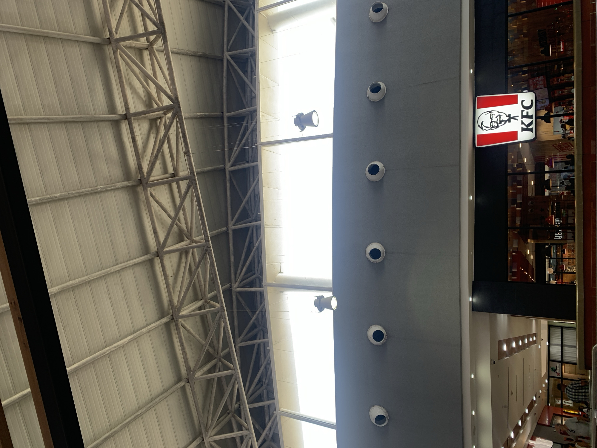 The image shows an upward view inside a building in Porlamar, Venezuela, featuring a KFC restaurant. The interior architecture includes a metal framework ceiling and a long vertical grey structure with recessed lights. A KFC sign is visible adjacent to a glass facade reflecting the interior of the restaurant. The area is brightly lit.