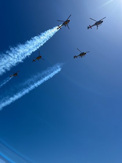 Five helicopters fly in formation against a cloudless, bright blue sky, leaving white smoke trails behind them. The sky occupies most of the frame. The bright sun creates a glare at the top right of the frame. The helicopters appear to be AgustaWestland AW109, likely HA.15. The scene is probably the city of Jerez de la Frontera, Spain during a military parade, possibly to celebrate the Day of the Armed Forces or similar.