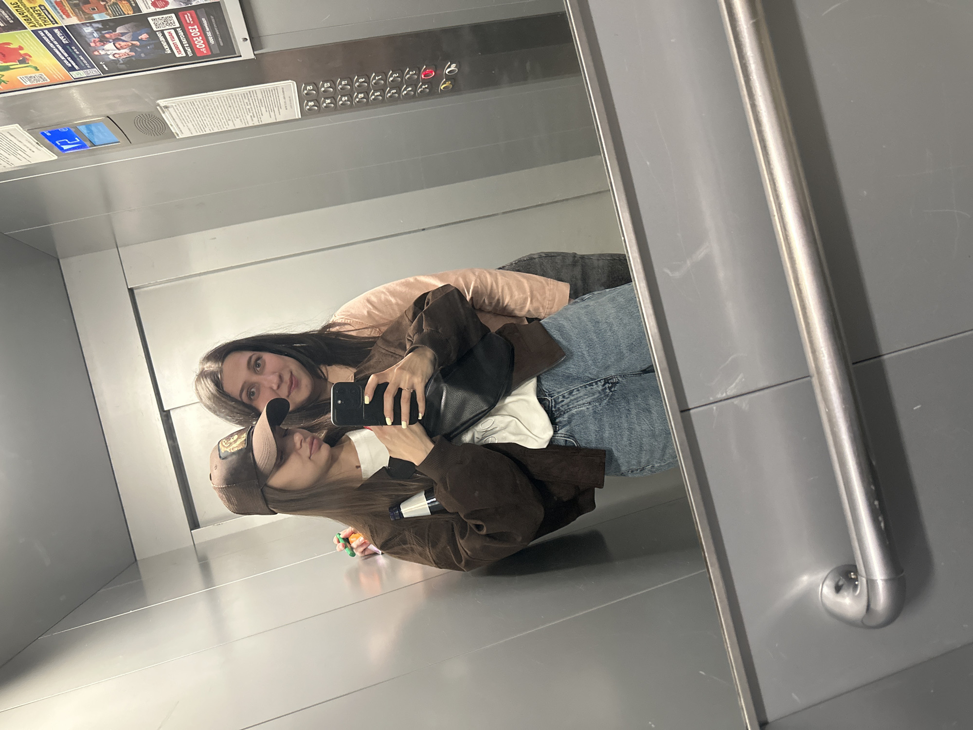 Two women are posing for a selfie in what appears to be an elevator. The woman in the foreground wears a brown baseball cap and a brown jacket. The other woman has long dark hair and wears a light pink top. They both hold a phone in their hands.
The elevator has gray walls and a control panel with buttons and signage. A promotional poster is visible at the top of the elevator, with text in Russian, including a price "130 200". There is also text that states "NHLBAU", possibly for a company or event. Given the presence of Russian text and the city is Izhevsk, Russia, this photograph was taken in that city.