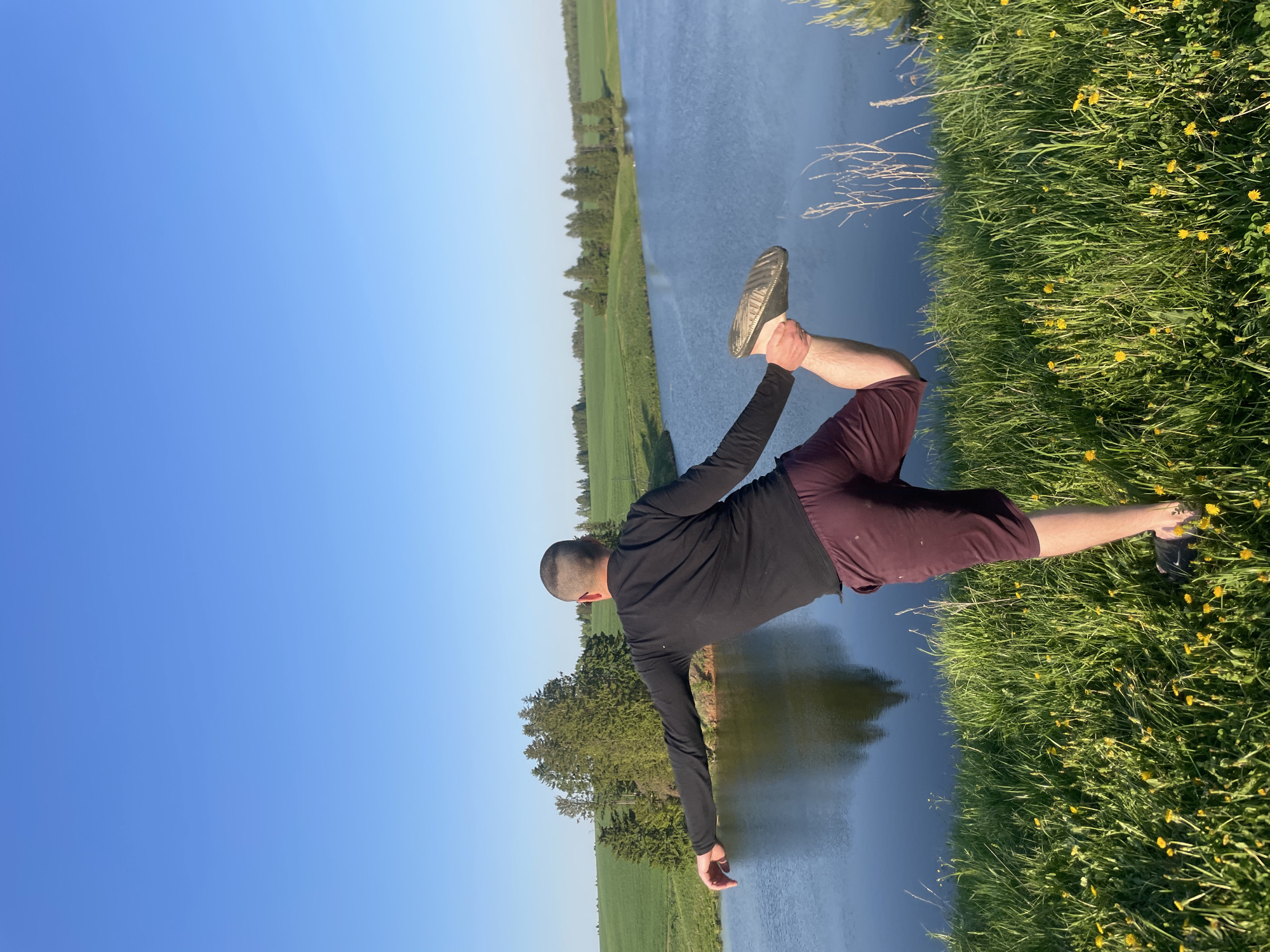A man is posing in a yoga-like pose on the bank of a body of water, likely a river or lake, in Подшивалово, Russia. He is wearing a black long-sleeved shirt and maroon shorts, with sneakers on his feet. He is standing on a grassy bank with yellow flowers. The water is to his left, reflecting the sky and the trees on the opposite bank. The sky is a clear blue. The man is holding one leg up behind him, grabbing his foot with his hand. The setting appears to be rural, with fields and trees in the background.