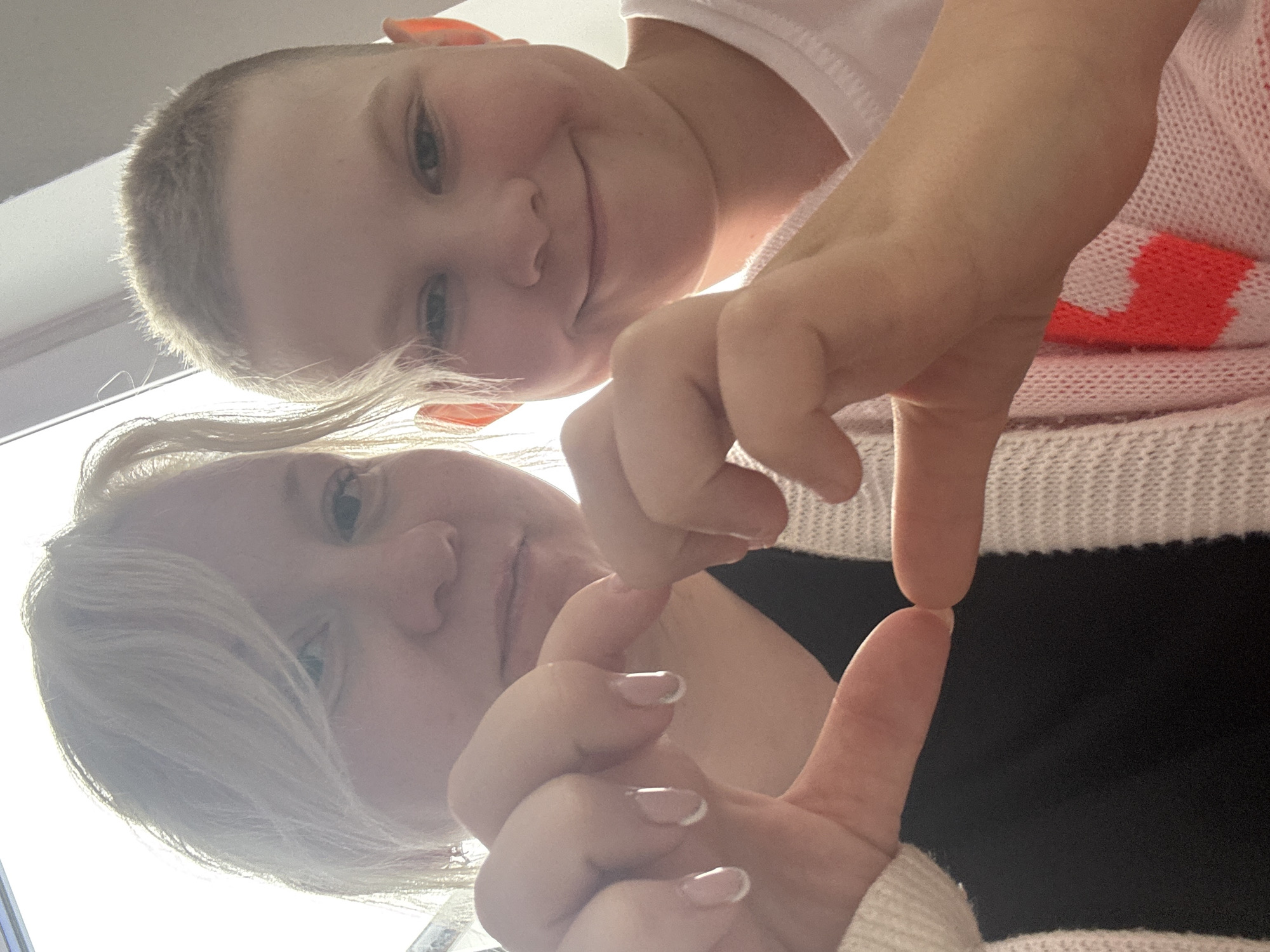A close-up, downward-angled selfie features two fair-skinned people. The top person, a child with short blonde hair, is smiling towards the camera. The bottom person has short blonde hair, possibly a woman, also looking at the camera. Both people appear to be interacting, with the person below making a hand gesture of a square, touching the index fingers and thumbs together. The hand partially obscures their face. The child wears a white shirt, while the other person is wearing a black top with a white and red patterned sweater over it. The background is bright. The exact location within Chaikovsky, Russia cannot be determined from the image.