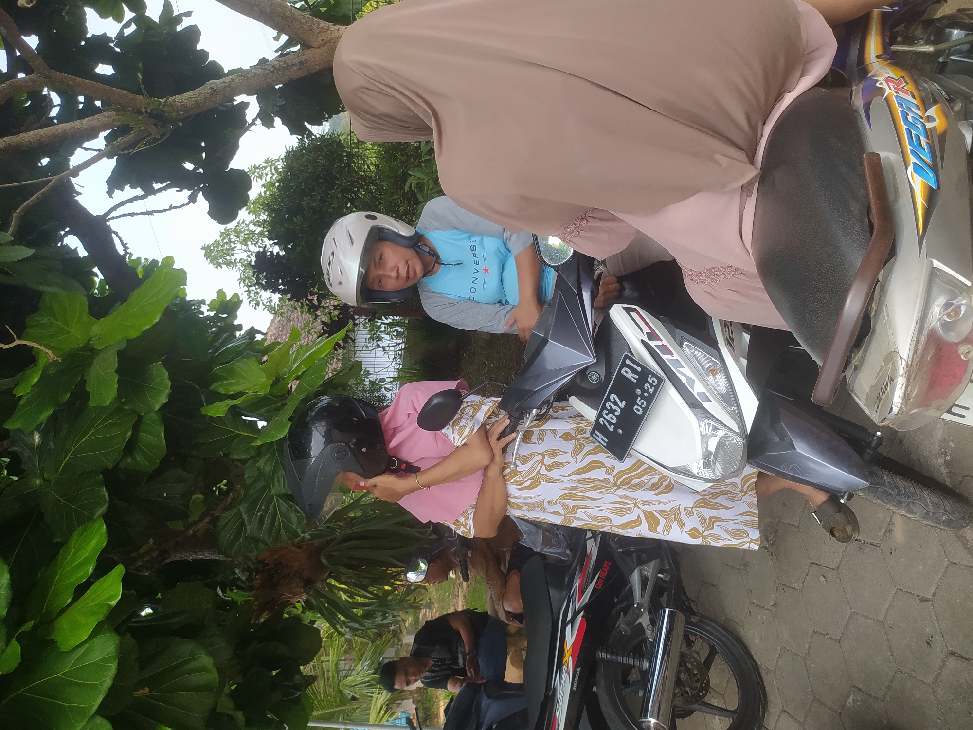 In Candirejo, Indonesia, three individuals are gathered outdoors near parked motorcycles. A woman in a pink hijab and a patterned dress stands next to a white motorcycle, adjusting her black helmet. To her right, another woman wearing a white helmet and a blue and gray shirt is seated on a motorcycle. In the background, two men are visible near other motorcycles, with one seated and the other standing. The scene is framed by lush green foliage and trees, suggesting a tropical environment.