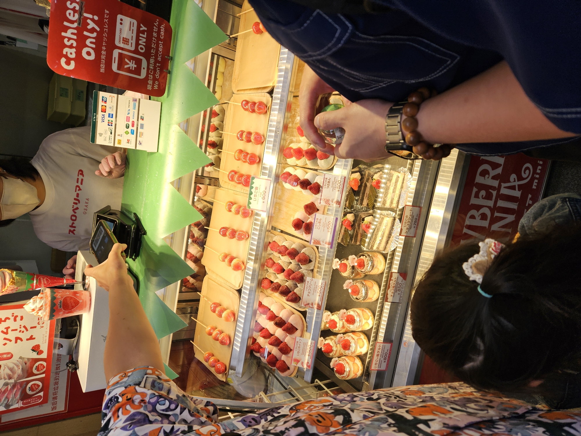 Here is a detailed description of the image:
**Scene:**
The image captures a scene inside a store, potentially in Osaka, Japan. It appears to be a sweets shop specializing in strawberry-themed desserts. The focus is on a display case filled with various strawberry treats.
**People:**
* **Employee:** There's a glimpse of an employee standing behind the counter wearing a white shirt with "ストロベリーマニア OSAKA" (Strawberry Mania Osaka) printed on it and a protective face mask.
* **Customer(s):** Several customers are visible, including a person taking a photo with their phone and a child with a hair accessory looking at the desserts. Another customer, wearing a watch and a bracelet, appears to be examining the products in the display case.
**Objects:**
* **Desserts:** The primary objects are the various strawberry desserts displayed in the case. These include strawberry candies on sticks, mochi with strawberries, strawberry whipped cream cups, parfaits, bottled strawberry tarts, and chocolate strawberry shortcakes. Prices are displayed for many of the items.
* **Display Case:** The desserts are arranged in a refrigerated display case with multiple tiers.
* **Cashless Payment Sign:** A prominent red sign reads "Cashless Only!" in both English and Japanese. It indicates that the store does not accept cash and displays logos for various credit cards and digital payment options.
* **Payment Terminal:** A smartphone and payment terminal are on the counter, ready for a cashless transaction.
* **Advertisement:** There's a sign or advertisement for strawberry parfaits.
**Actions/Interactions:**
* **Shopping:** Customers are browsing and selecting desserts.
* **Payment:** One person is in the process of paying with their mobile phone.
* **Photography:** A customer is taking a photo of the desserts with their smartphone.
**Environment:**
* **Bright Lighting:** The display case is well-lit to showcase the desserts.
* **Clean and Organized:** The display case appears clean and well-organized, with items arranged neatly.