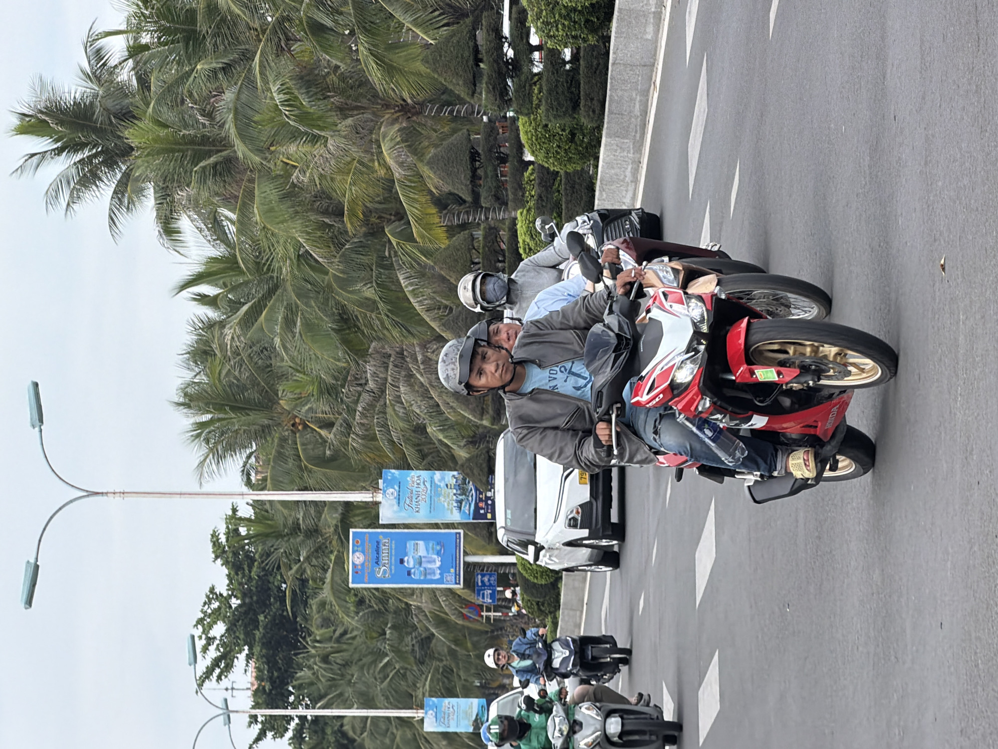 The image depicts a street scene in Nha Trang, Vietnam, with multiple people riding motorcycles. In the foreground, a man wearing a gray jacket and a helmet is riding a red and white motorcycle. To his left, another man with a helmet is riding a motorcycle, and to the left of him, a person in a green jacket and helmet is also on a motorcycle. In the background, several other motorcycles are visible, along with cars and a white SUV. Palm trees line the street, and several banners with text and images are displayed on poles. The sky is overcast. The overall impression is of a busy street with common modes of transportation in Vietnam.