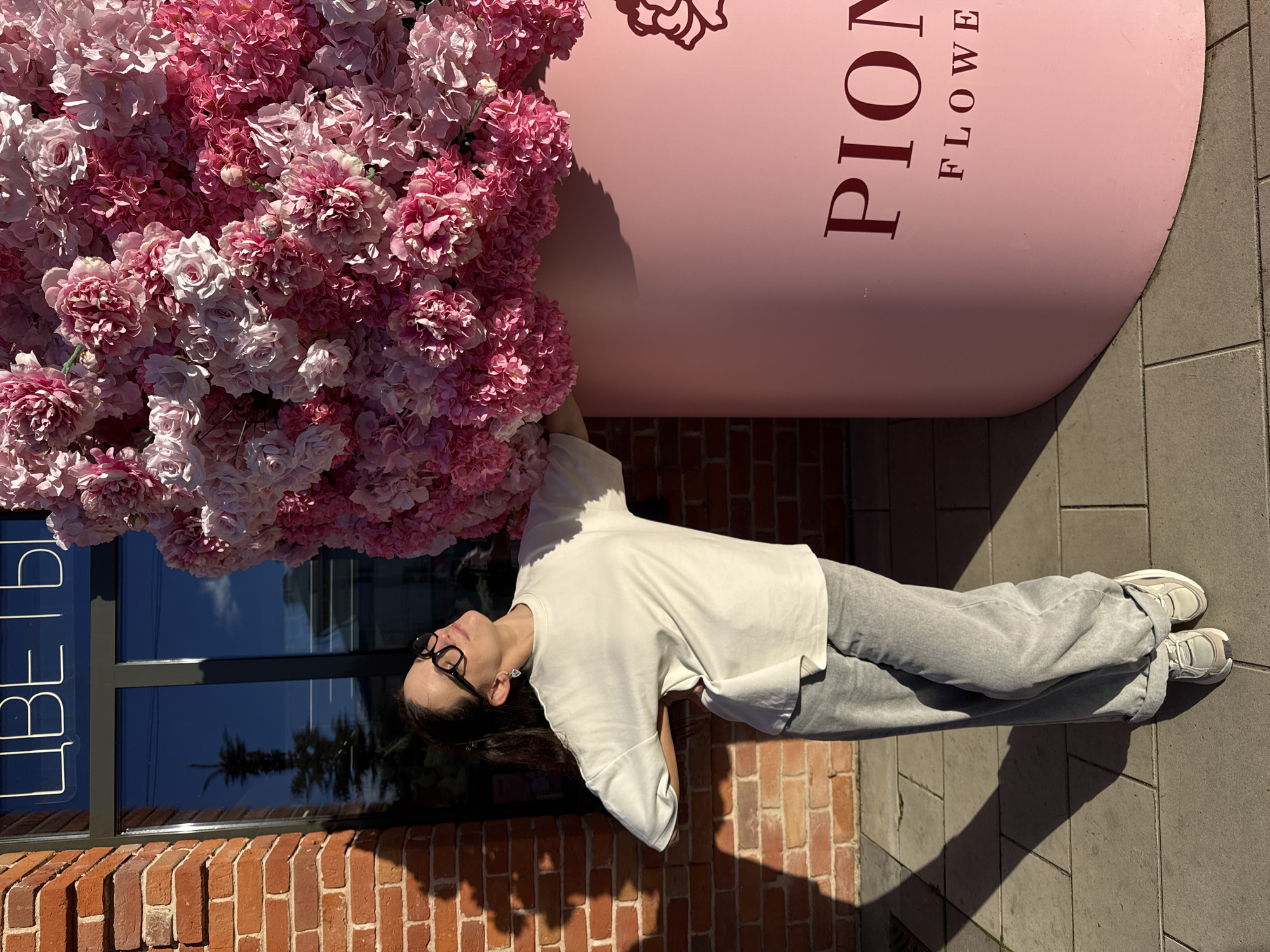 A woman in a white t-shirt and grey pants stands next to a large pink cylindrical planter overflowing with pink and white artificial flowers. She is looking up and to the right with her eyes closed and her left hand on her lower back. The planter has the word "PION" and "FLOWERS" written on it in dark red lettering. Behind the planter is a brick building with a window that has the word "ЦВЕТЫ" (which means "Flowers" in Russian) written in neon white letters. The scene is outdoors on a paved walkway, likely in the city of Kazan, Russia, given the Russian text on the building. The sun is casting shadows on the ground and the brick wall.