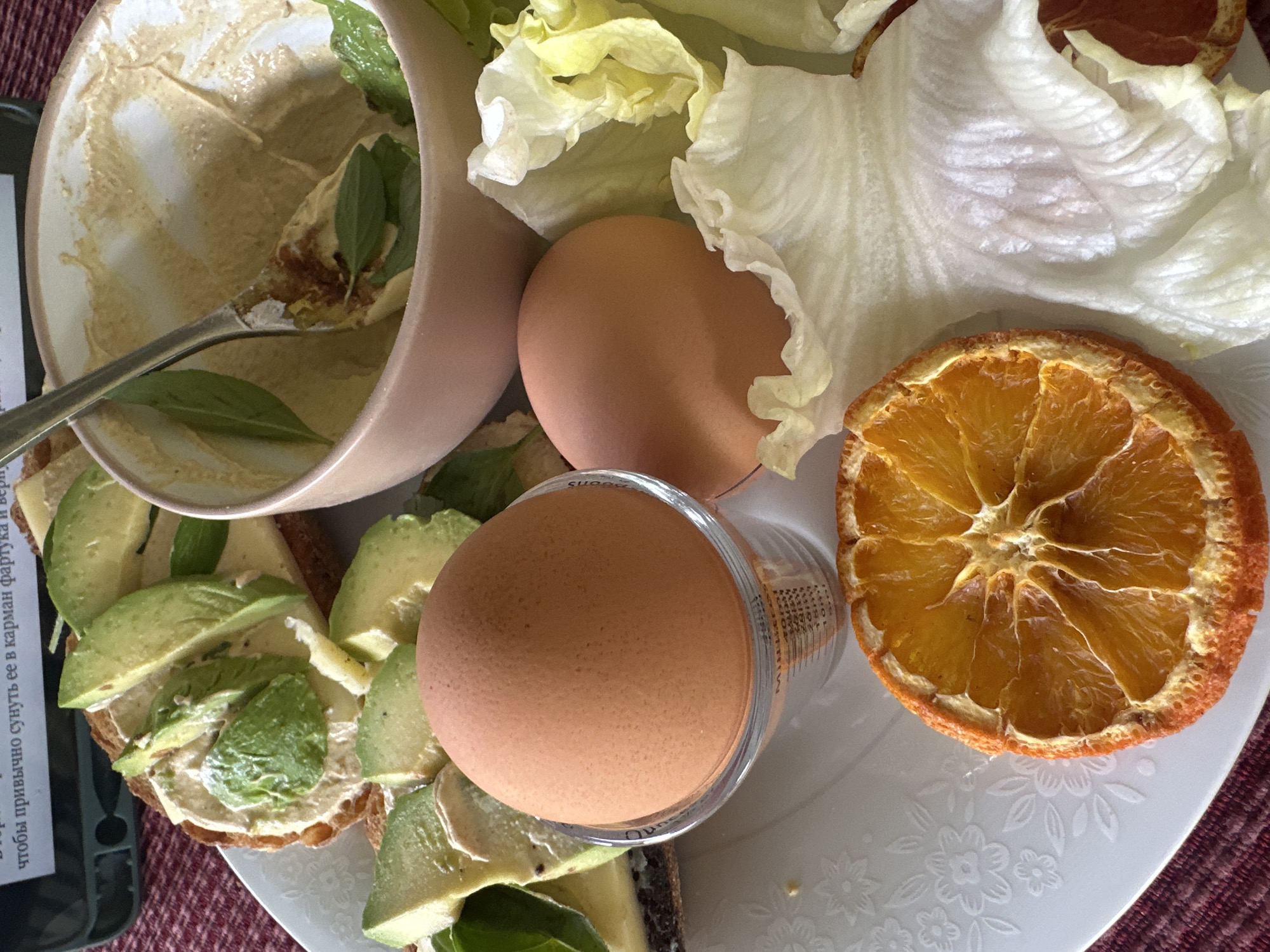 The image shows a close-up of a breakfast plate. On the plate, there are two hard-boiled eggs, one in a clear measuring cup, sliced avocado on toast with cheese and greens, a bowl with some remaining dip or spread and a spoon, lettuce leaves, and a dried orange slice. The plate is on a maroon and white checkered tablecloth. There is text in Russian on a dark green bottle in the background. The image does not provide any information about the location being in Thora, Australia.