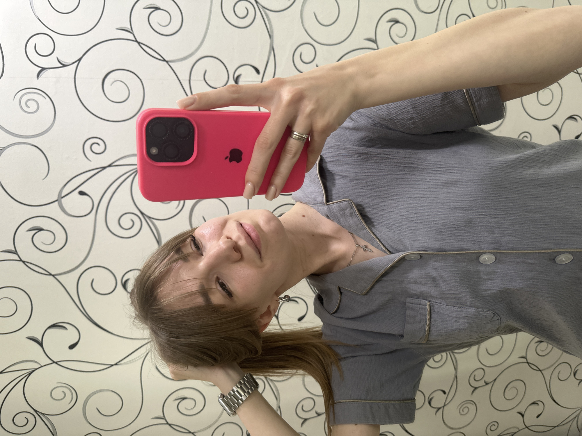 A woman is taking a selfie with a pink smartphone. She is wearing a gray collared shirt and a silver ring on her finger. She also has a silver watch on her wrist. Her hair is brown. The background is a wallpaper with black and white swirl patterns. The location in the city of Chaikovsky, Russia is not discernable from this image.