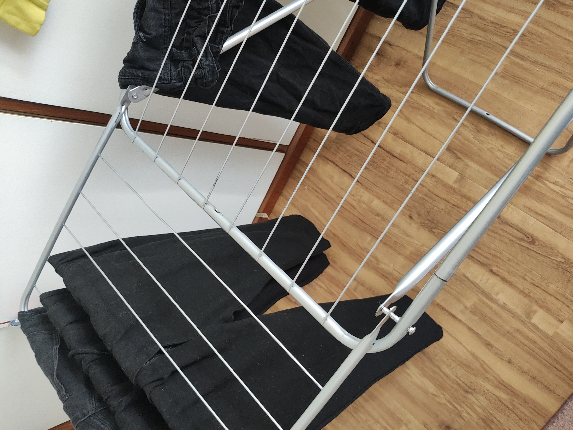 An indoor scene in Trebgast, Germany, depicts a silver-colored metal clothes drying rack placed on a light brown wooden laminate floor. Four pairs of dark-colored trousers or jeans are draped over the rack's bars, with three pairs occupying the left side and one pair on the right. The background features a white wall or cabinet sectioned by a vertical dark brown wooden trim. A yellow object is partially visible in the upper right. No persons are present.