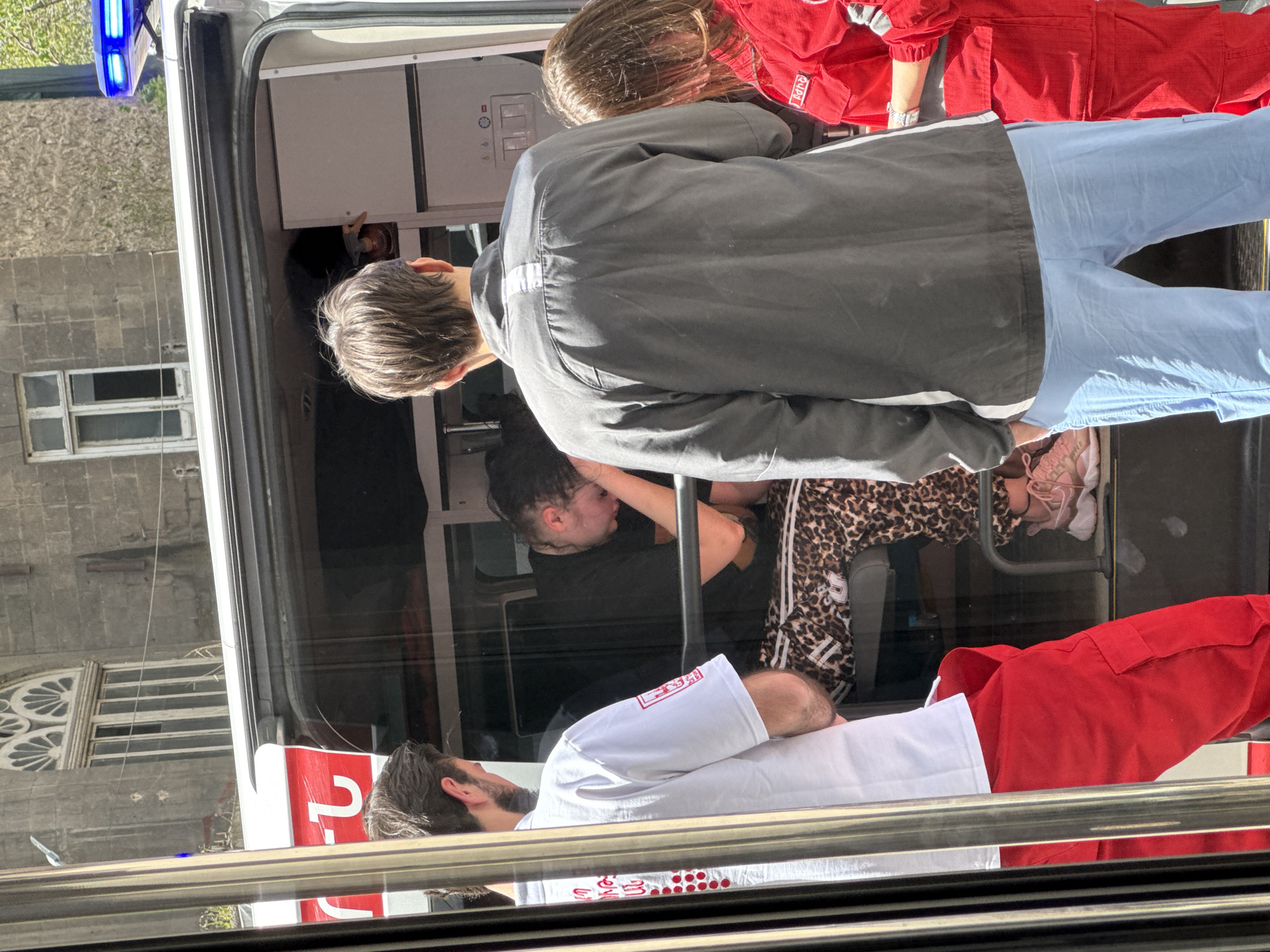 Captured from outside, the image depicts the interior of what appears to be an ambulance in Yerevan, Armenia. Inside the vehicle, a person is lying down, being attended to by two individuals wearing medical uniforms with a red and white cross insignia. One medic is dressed in a red jumpsuit with white details, while the other wears a white shirt over red pants. Another person in casual attire is leaning over the patient. The background features a building with distinctive window architecture and the blue lights of the ambulance. The scene suggests a medical emergency.