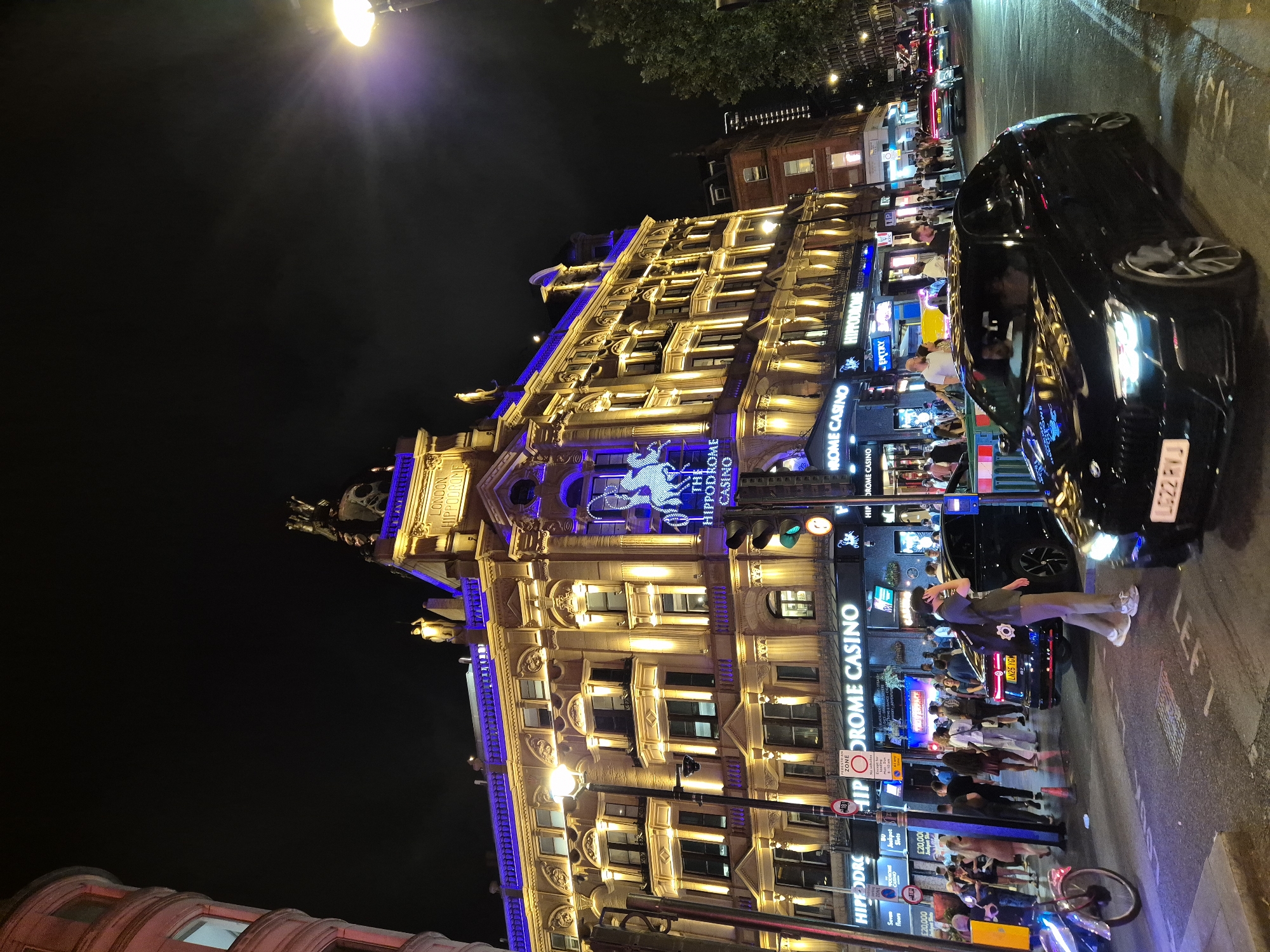 The image captures a vibrant nighttime scene in London, United Kingdom, focusing on the iconic London Hippodrome Casino. The grand building, illuminated with warm yellow lights and accented with a striking blue hue along its upper edges, dominates the frame. The casino's name is prominently displayed in illuminated signs. In the foreground, a busy street scene unfolds. Several black cars are present, with one in the immediate foreground appearing to be a modern luxury sedan with its headlights on. Pedestrians are visible on the sidewalks and crossing the street, suggesting a lively atmosphere. Traffic lights are also visible, indicating an active intersection. The overall impression is one of a bustling urban environment at night, characterized by bright lights, architectural grandeur, and the presence of people and vehicles.