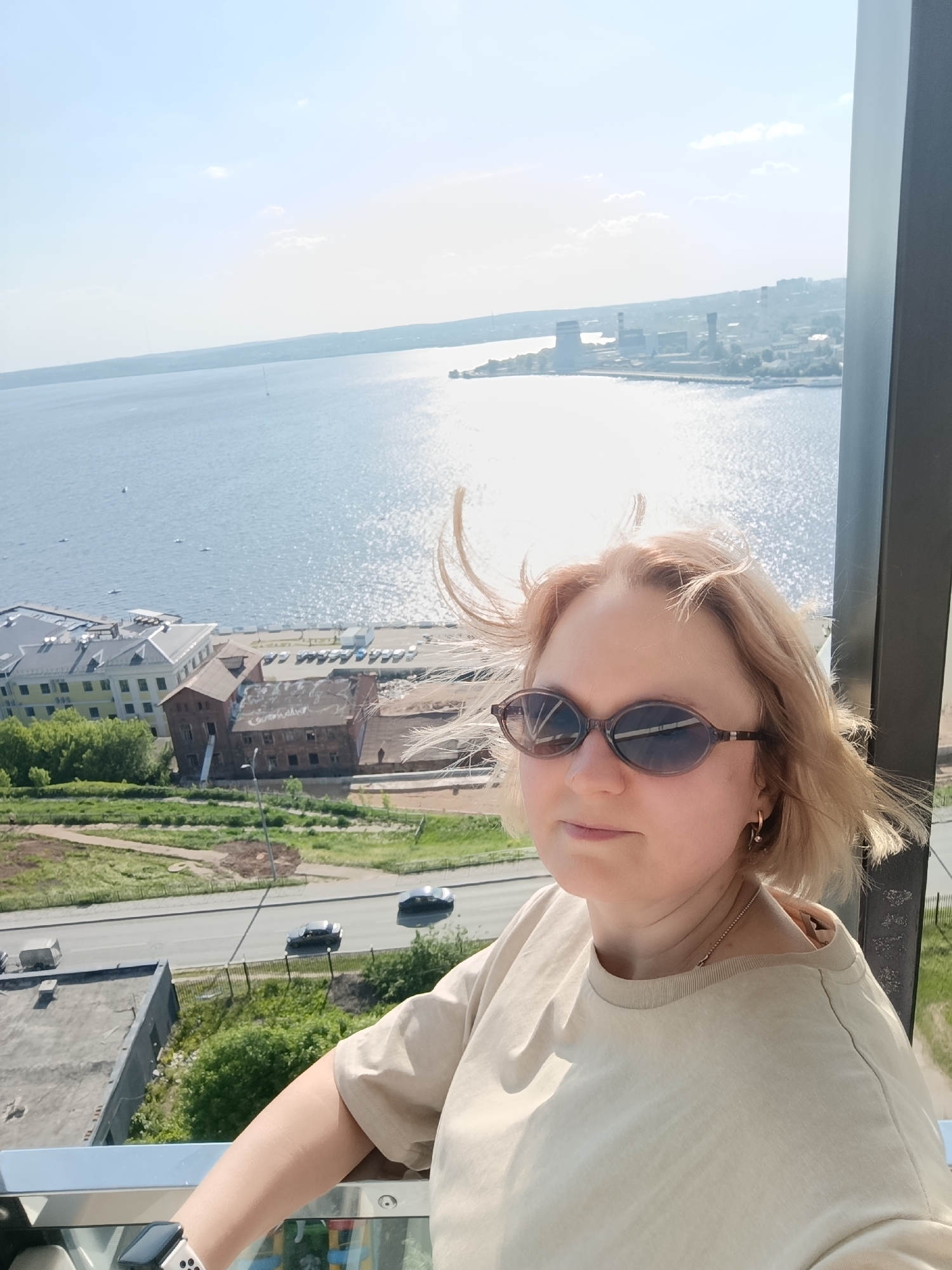 A woman is standing on an observation deck in Izhevsk, Russia, overlooking a cityscape and a body of water. She is wearing sunglasses and a beige t-shirt. Her blonde hair is being blown by the wind. In the background, there is a river with boats, a city skyline, and several buildings. There is a road with cars below. Some of the buildings in the foreground are historical with red brick and grey roofs. A modern glass panel and metal support is on the right side of the image.