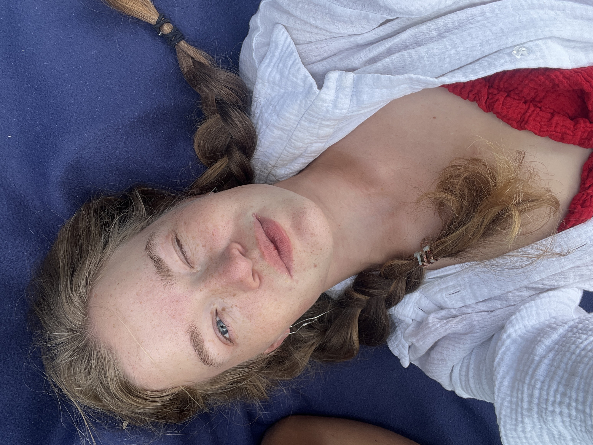 A high-angle, portrait-style photograph shows a fair-skinned individual, presumed to be female, lying on a blue textured surface. She has light brown hair styled into two braids, secured with dark and light-colored bands. Her eyes are blue, and her face is freckled. She is wearing a white, textured shirt, partially unbuttoned, revealing a red garment underneath. The setting is ambiguous and there are no indicators to the location of Velji Bostur, Montenegro.