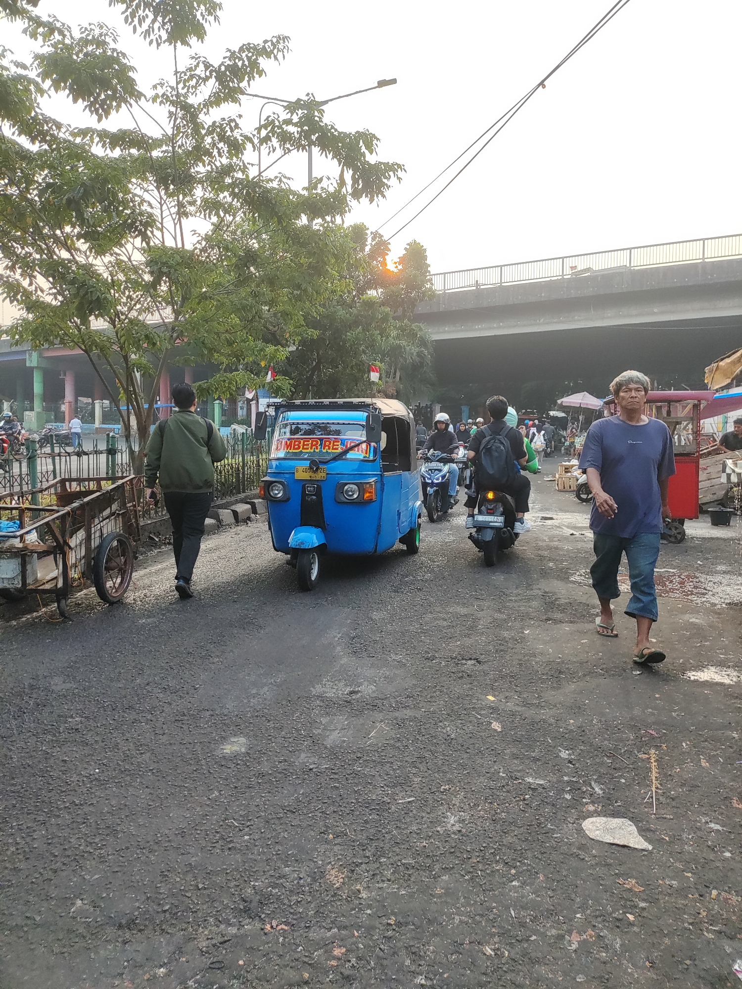 In East Jakarta, Indonesia, an outdoor scene unfolds along a paved road. The foreground is dominated by a blue, three-wheeled auto-rickshaw, also known as a "bajaj," with the text "AMBER REKI" visible on its windshield. To the left of the auto-rickshaw, a man in a green jacket walks away from the camera, carrying a dark bag. Beside him is a rusty, two-wheeled cart.
Further down the road, a man in a dark blue t-shirt and denim shorts walks towards the viewer. Behind him, a motorcycle carries two individuals, the driver wearing a helmet and the passenger with a black backpack. In the background, an elevated concrete bridge crosses overhead, its structure supported by pillars. The sky is overcast, with hints of sunlight peeking through the clouds. Trees with green foliage line the sides of the road and the area beneath the bridge. A market scene is suggested by the presence of stalls and awnings to the right of the frame. The road surface is dark and appears somewhat uneven, with scattered debris. The overall impression is of a bustling urban environment during daylight hours.
