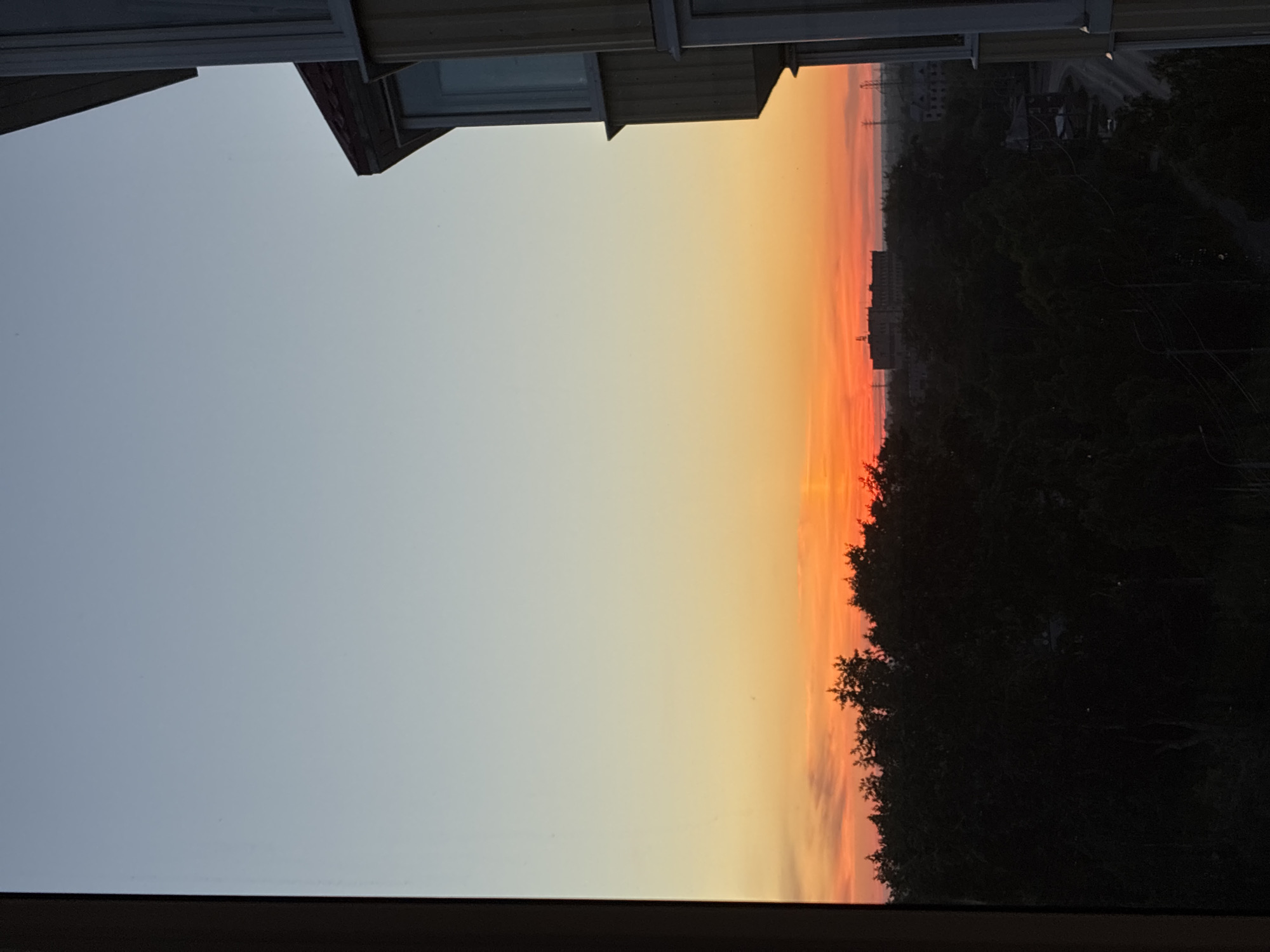 An outdoor, vertical, eye-level medium shot shows a view from a window of a sunset over a densely wooded area. The sky is a gradient from a pale blue at the top to a bright orange with some pink clouds near the horizon. The sun is a small, bright orange disc low on the horizon, partially obscured by the trees. The trees form a dark, silhouetted mass across the lower third of the image. In the middle distance, there are several buildings, including a large, elongated building with multiple windows and a flat roof, as well as some smaller structures. The foreground is dominated by the dark frame of the window and the side of a building with yellow siding and vertical white trim. The overall impression is one of a peaceful, end-of-day scene.