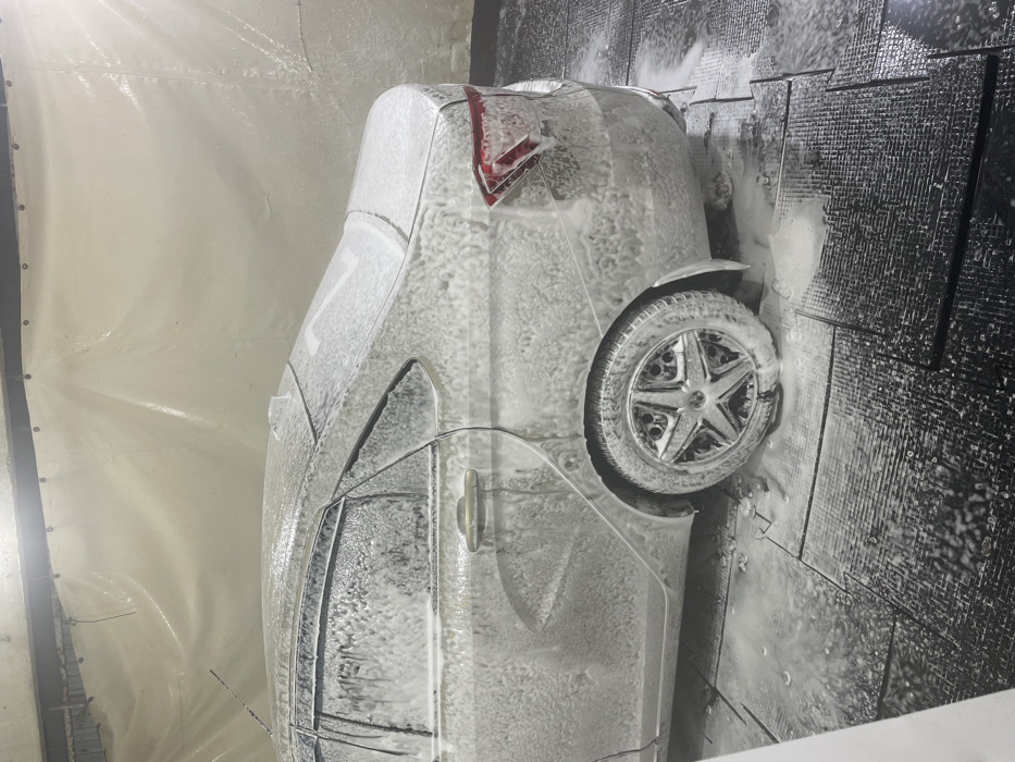 A silver sedan, likely a Lada Vesta, is positioned inside a car wash bay in Zubovka, Russia. The vehicle is completely coated in a thick layer of white soap foam, indicating it is undergoing a wash cycle. The car is oriented in a rear three-quarter view from the driver's side, showcasing the rear passenger door, rear wheel with a five-spoke alloy rim, and the rear tail light assembly, all obscured by foam. Water and soap residue are visibly accumulating on the dark, textured, interlocking rubber or plastic floor tiles beneath and around the vehicle. The background consists of light-colored, possibly translucent, plastic or tarp curtains suspended from an overhead metal beam, typical of an enclosed car wash facility. Artificial overhead lighting illuminates the scene. No individuals are present in the frame.