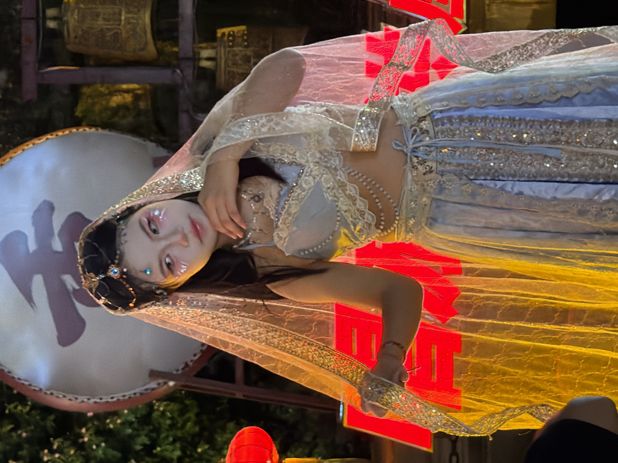In Yuzhong District, China, a young woman stands adorned in traditional Chinese attire, her head covered by an intricately embroidered veil. She wears a light blue, embellished bra-like top and a flowing, light blue skirt that shimmers with sequins. Her makeup is theatrical, featuring glitter on her cheeks and stylized eye makeup. She holds a piece of the veil in her hand, which is adorned with a jeweled headband. In the background, a large drum with Chinese calligraphy, a display of brass bells, and red lanterns are visible. The lighting is dramatic, with bright red light illuminating the lower portion of the scene, creating a festive and somewhat mysterious atmosphere.