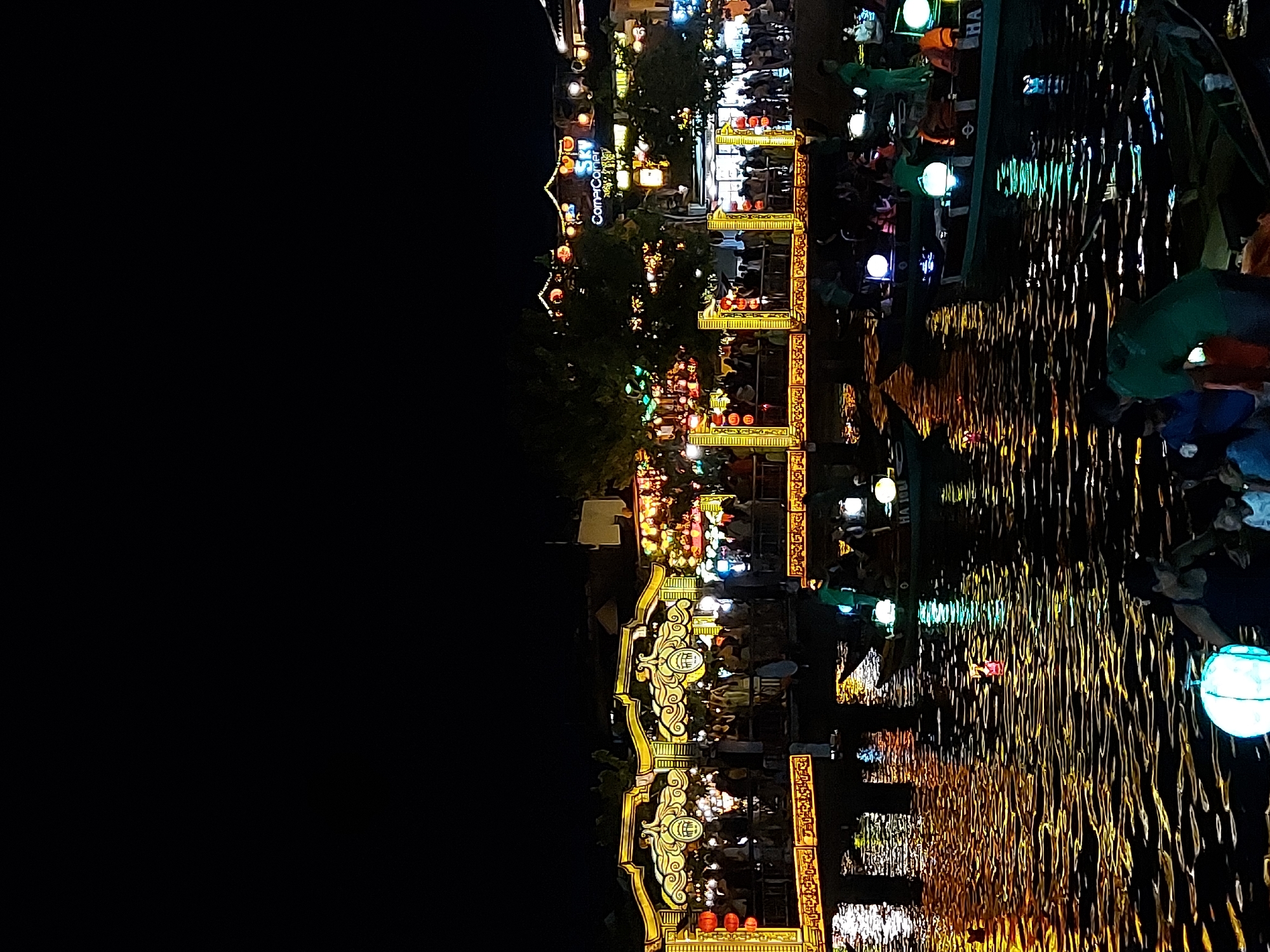 The image depicts a nighttime scene along the Thu Bon River in Hoi An, Vietnam, with the iconic Japanese Covered Bridge brightly illuminated in the background. The bridge, a prominent landmark, is adorned with yellow decorative lights and lanterns, with many people gathered on and around it. Several boats are on the river, some with passengers, and many are decorated with glowing lanterns. The water reflects the vibrant lights from the bridge and the surrounding buildings, creating a festive and bustling atmosphere. In the foreground, people are visible near the water's edge, some interacting with lanterns. The overall scene is characterized by a celebration involving lights and water, indicative of a festival or a popular tourist activity in Hoi An.