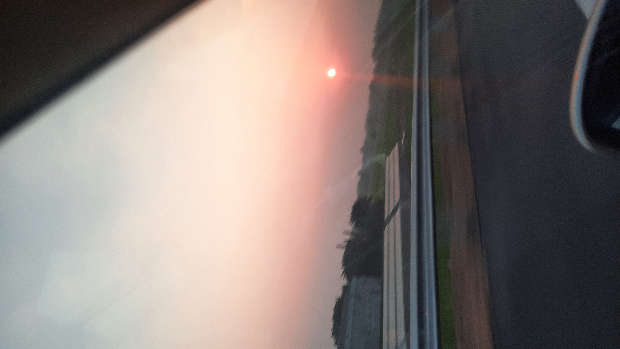 The image depicts a blurry, low-resolution view of a sunset over a rural landscape, likely taken from inside a moving vehicle. The sky is a gradient of pale blue to pink, with the sun appearing as a small, bright orb low on the horizon, casting a warm glow. In the foreground, a dark, possibly asphalt road is visible, with a metal barrier separating it from a strip of green vegetation. Beyond the vegetation, there are structures that appear to be greenhouses or agricultural buildings, along with trees and more vegetation. The overall impression is one of natural scenery viewed during twilight from a roadside perspective. The location in Sumuran, Indonesia, is not explicitly depicted in the image.