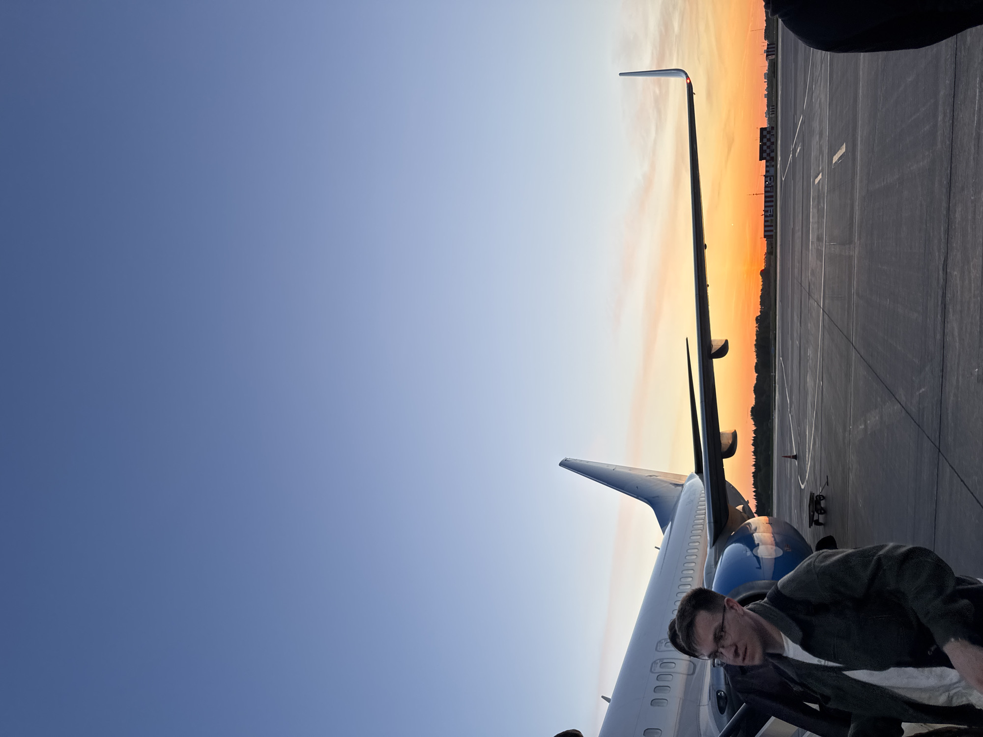 The image shows a man with glasses standing next to a passenger airplane. The airplane has a blue tail fin and a white body. The background shows an airport tarmac and a sunset. The photograph may have been taken at Kitsjanovo airport in Russia.