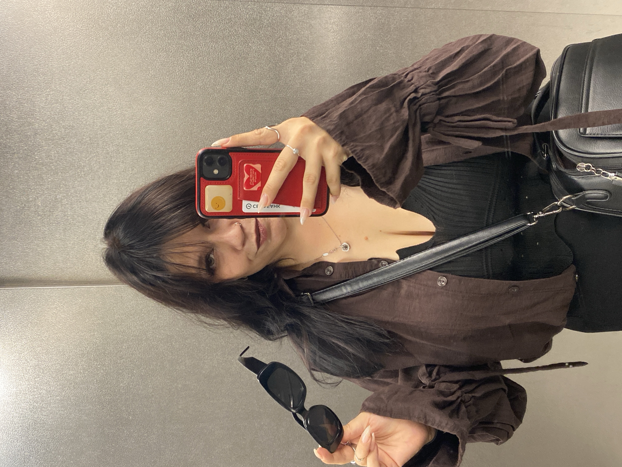 A young woman stands in an elevator, holding sunglasses in her left hand and her red smartphone in her right, taking a mirror selfie. She has dark, wavy hair and is wearing a dark brown, button-up blouse with ruffled sleeves over a black ribbed top, and black bottoms. She is accessorized with a silver necklace and rings. A black handbag is slung over her right shoulder. The elevator walls are a textured grey metallic material.