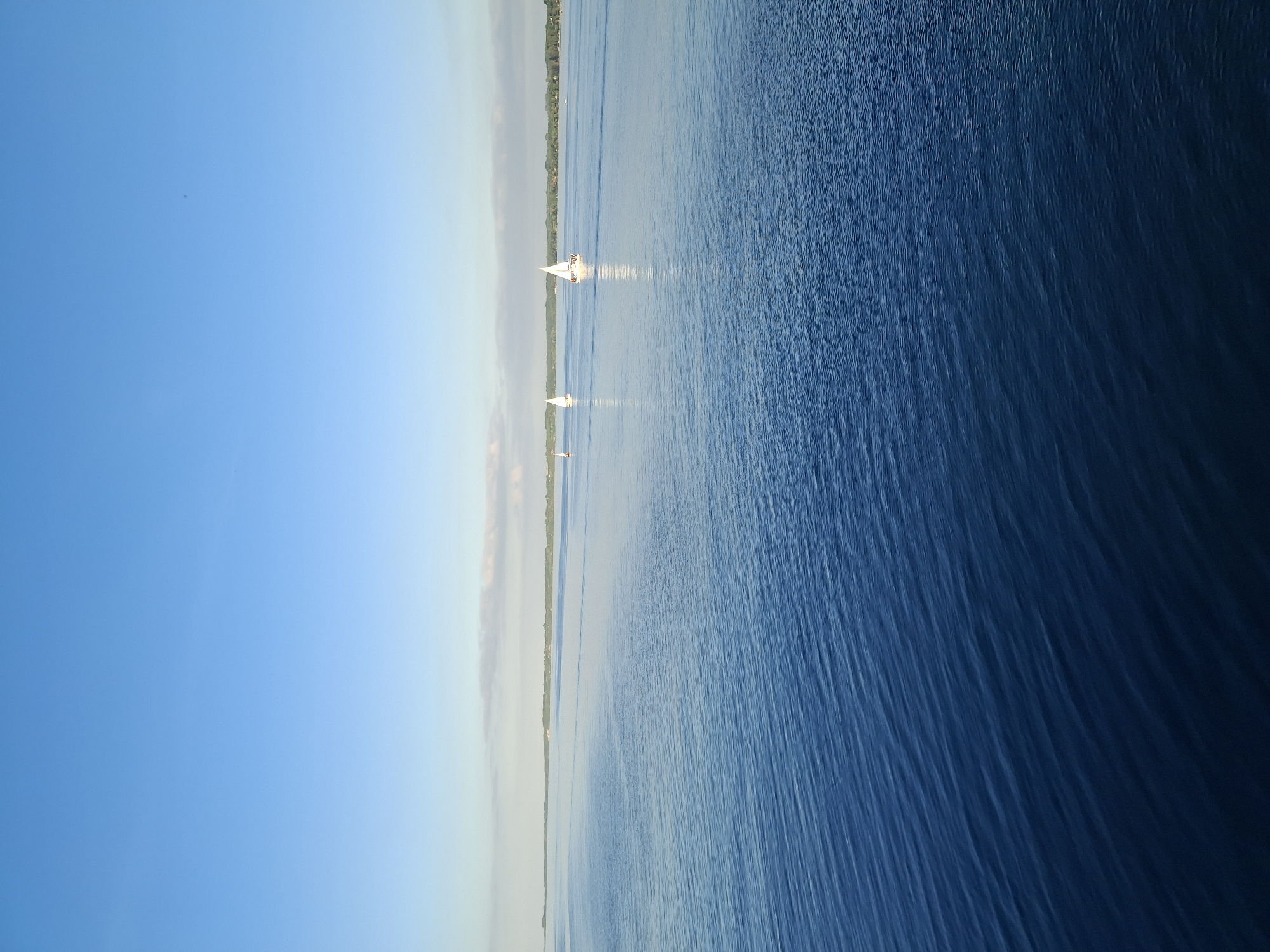 A landscape photograph shows a body of water, possibly a lake or a sea, with a narrow strip of land visible in the middle. The water transitions from lighter to darker shades of blue as it stretches from left to right. There are several sailboats with white sails visible on the water near the land, which appears to have trees along the shoreline. The sky on the left side of the image is a clear, light blue, contrasting with the darker hues of the water on the right. The photograph is taken from an elevated position, giving a panoramic view of the scene. I don't have information on the location, and thus cannot say anything about whether it is in Aabenraa, Denmark.