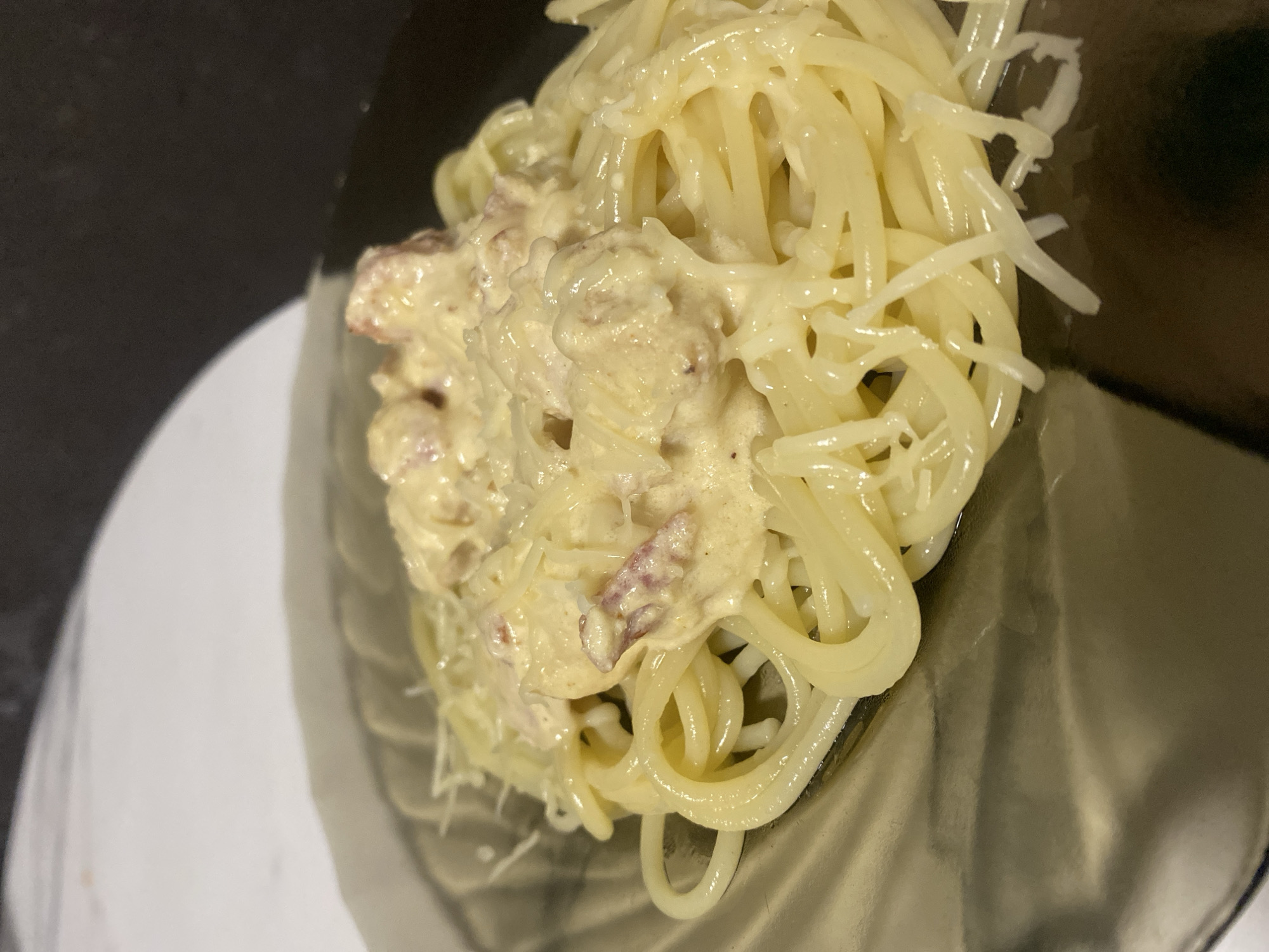 A close-up, slightly tilted, eye-level shot depicts a plate of spaghetti carbonara. The dish is presented on a transparent or smoked glass plate placed atop a white surface. The spaghetti noodles are coated in a creamy, light-colored sauce with visible pieces of what appears to be pancetta or bacon. A dusting of shredded cheese is scattered over the dish. The background is dark, providing a contrast to the creamy pasta. The location of the image is unknown.