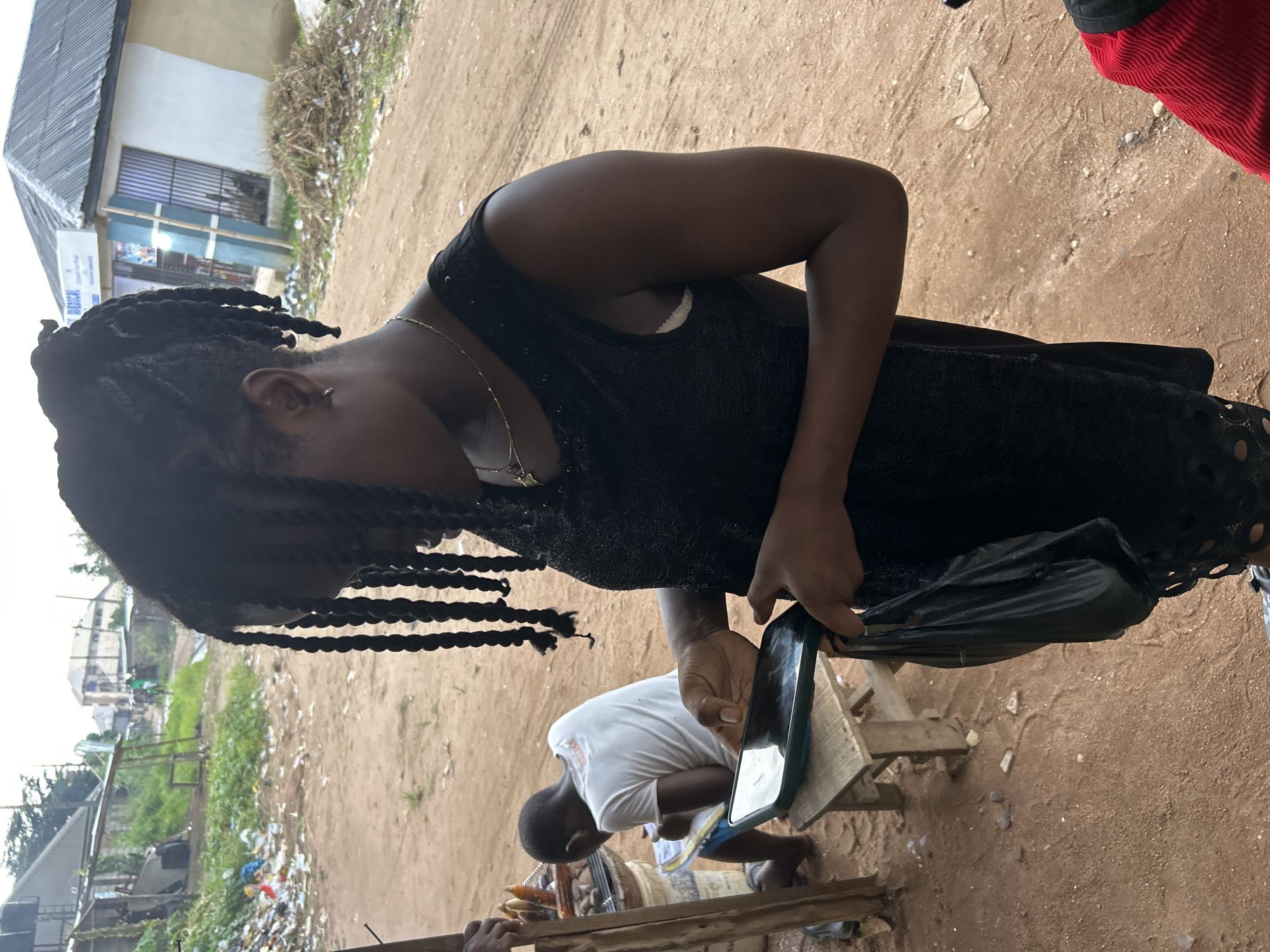 An outdoor medium shot captures a woman from the chest up, wearing a black sleeveless dress and a gold necklace. She is holding a smartphone in her left hand and a black plastic bag in her right hand. Her hair is styled in long braids that fall forward, obscuring her face. In the background, to the left of the woman, a young boy wearing a white t-shirt sits on a wooden stool, appearing to write in a notebook. To the far left, a wooden stall displays what seem to be ears of corn. The environment appears to be a dusty, unpaved street in a low-income area, possibly in Ihiagwa, Nigeria. In the background, there are buildings with corrugated metal roofs, piles of refuse, and overgrown vegetation, suggesting a rural or semi-urban setting. The lighting indicates it is daytime, with overcast skies.