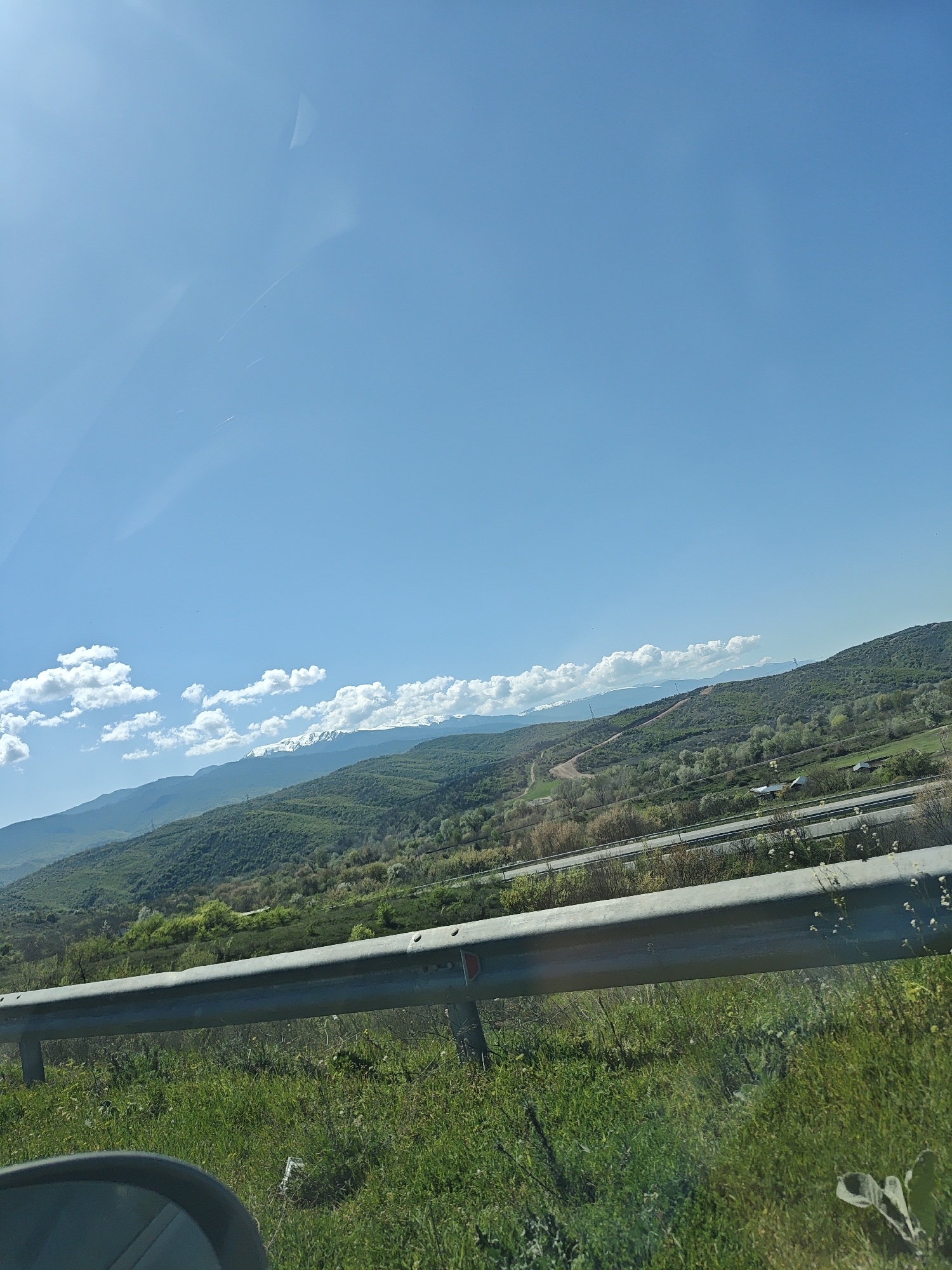 The image depicts a landscape with a clear blue sky and scattered white clouds. Mountains are visible in the background, some with snow-capped peaks. The foreground features green grass and a gray metal guardrail. A road is seen in the middle ground, running alongside the mountains. A portion of a side mirror is visible in the lower left corner. The location is identified as Unknown, Unknown.