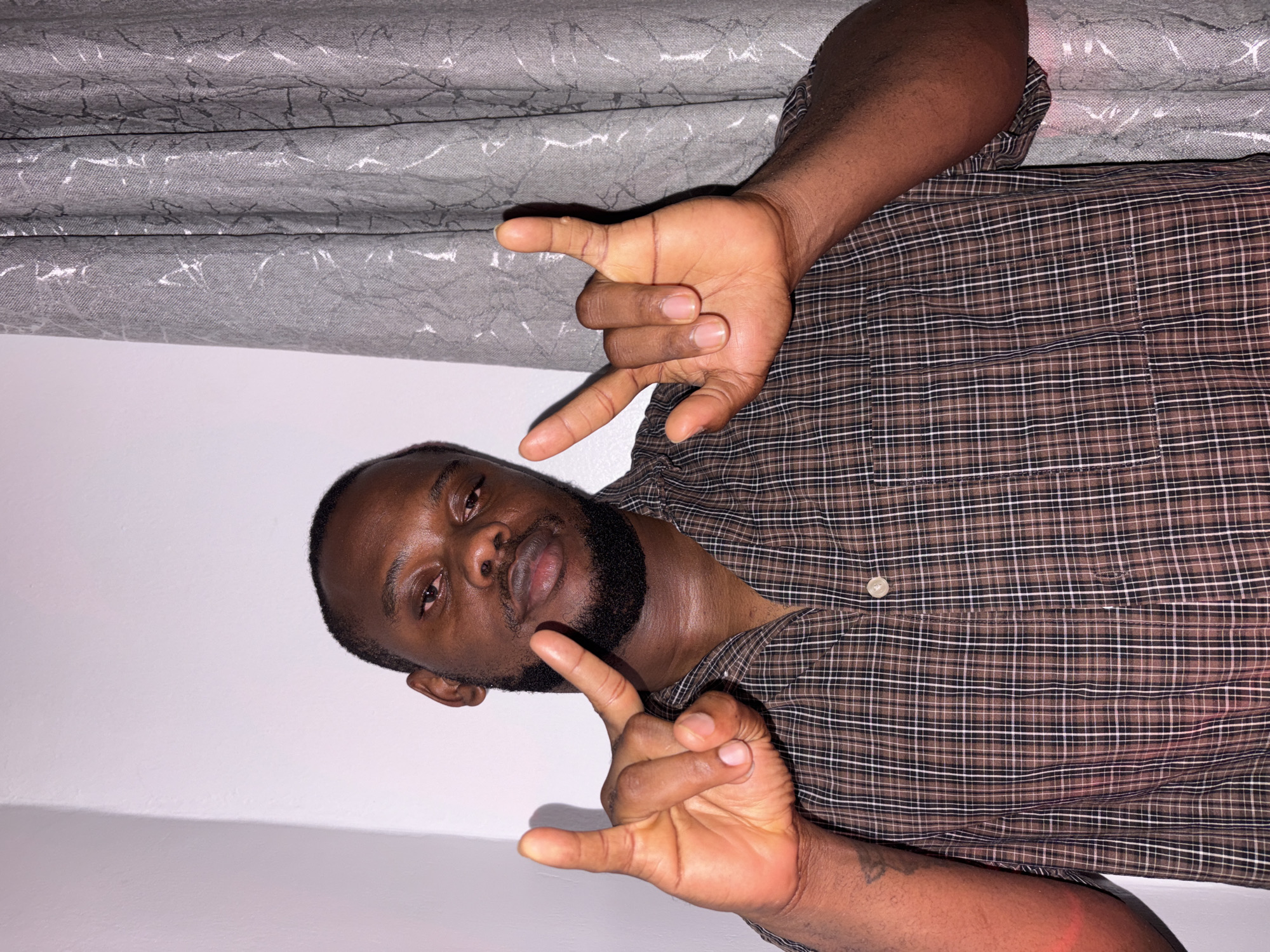 An adult Black man is posing for a photo. He is indoors, standing in front of a wall with a gray curtain or drape partially visible at the top of the frame. He is wearing a brown and black plaid button-up shirt. He is holding his hands up, fingers spread in a gesture resembling the "devil horns" or "rock on" hand sign. The location is unknown and not provided in the image. The details provided in the prompt about Iddo Tudunwada, Nigeria are also not provided.